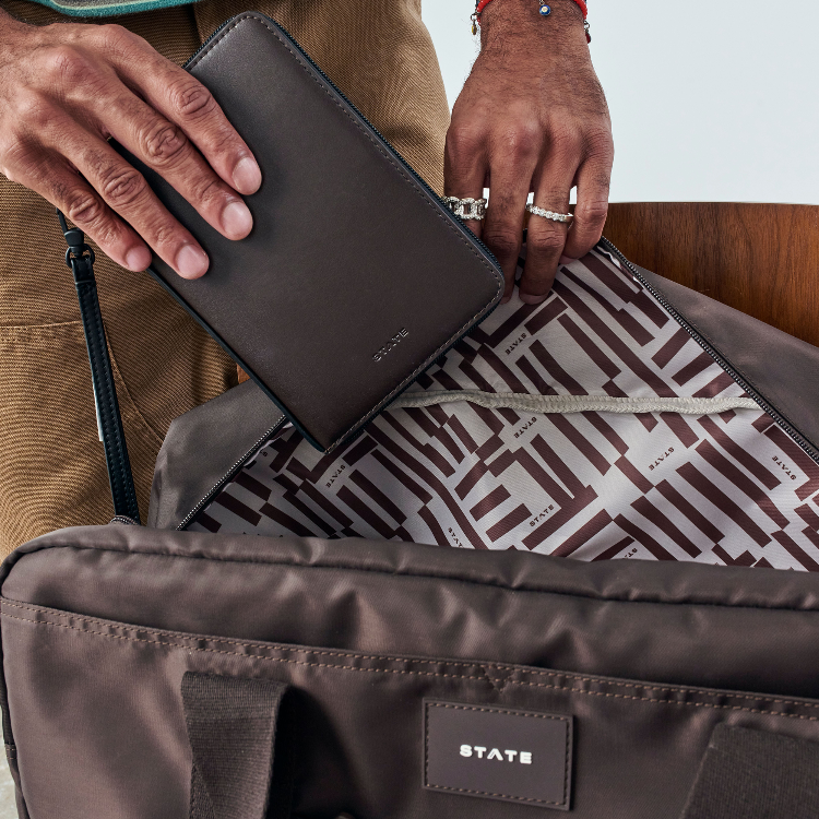 A close-up of a person's hands holding a dark brown leather wallet next to an open brown nylon bag with a patterned interior and a visible STATE logo patch.