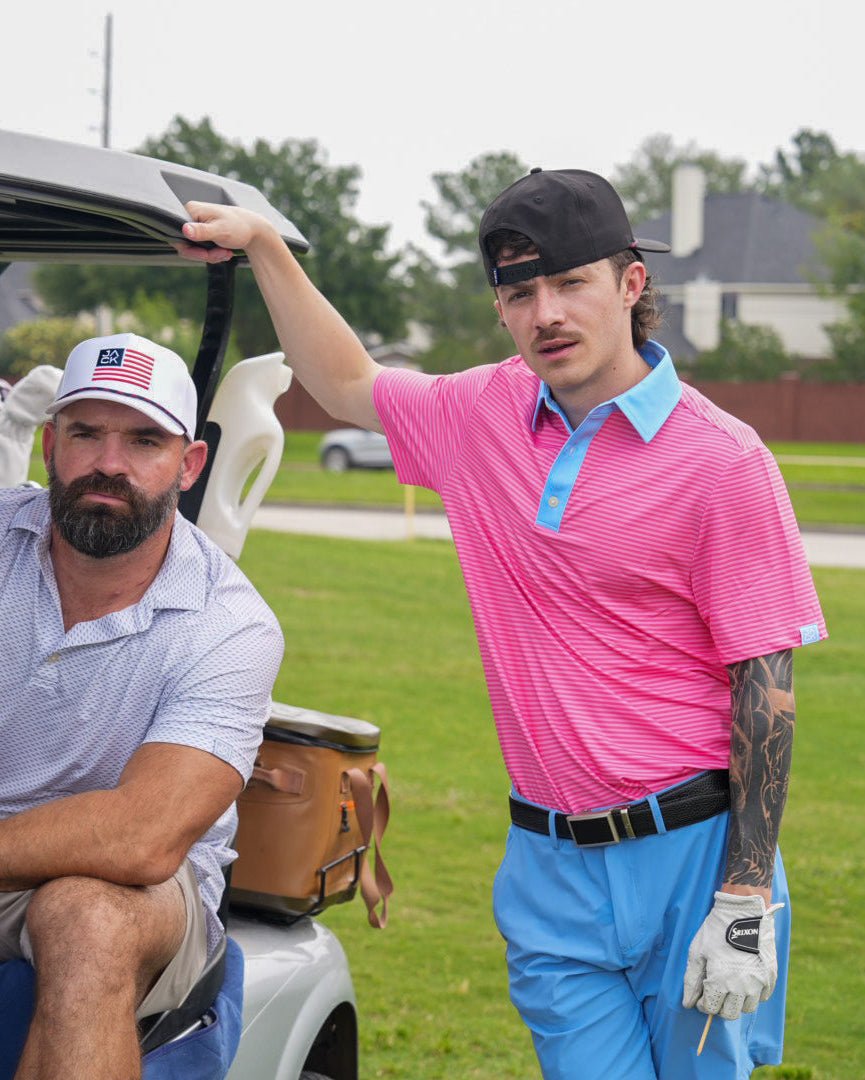 Two men on a golf course, one in a pink shirt and blue pants, the other in a white cap and light shirt, with a golf cart in the background.