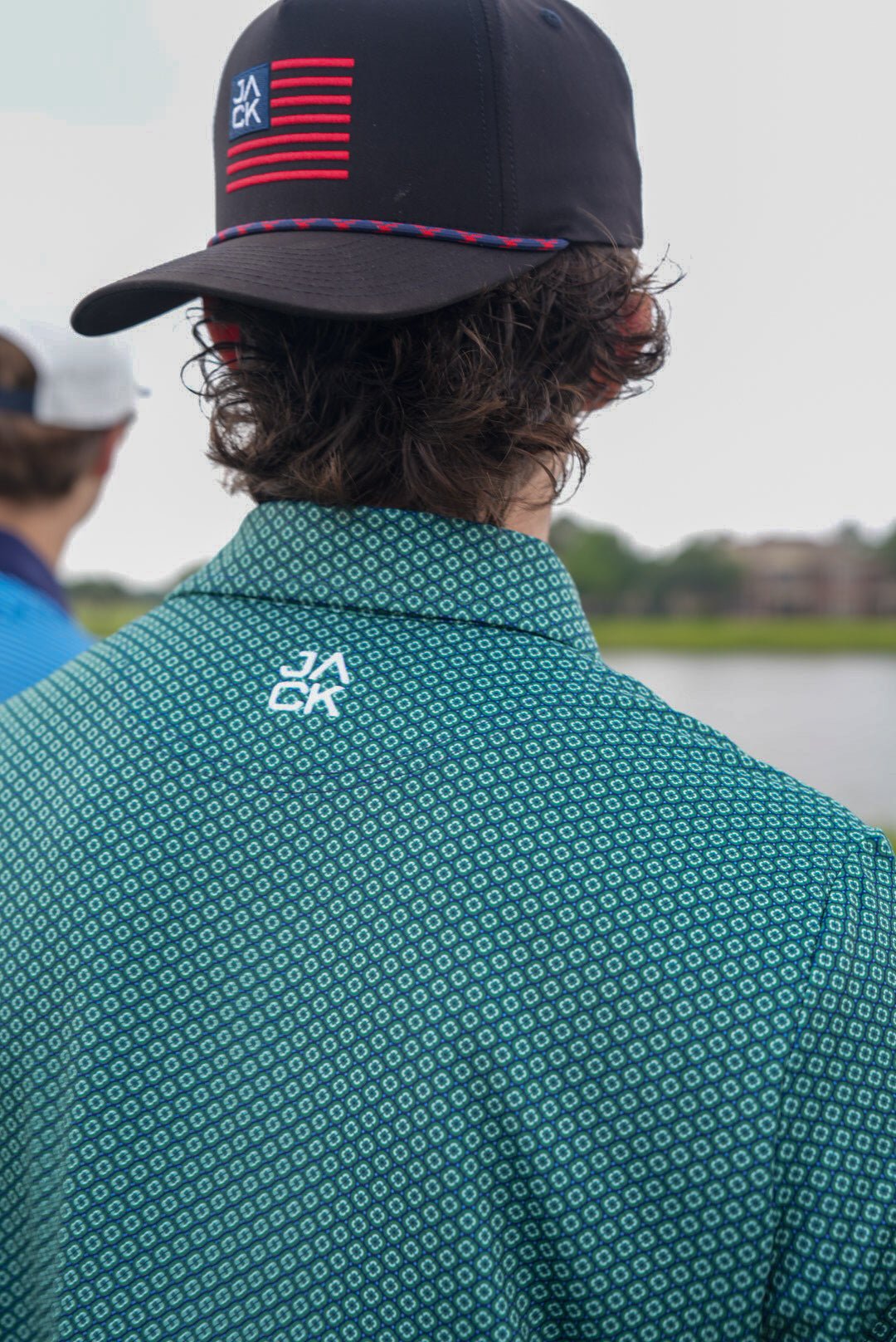 Person wearing a teal patterned shirt with a logo, looking over their shoulder towards a body of water.