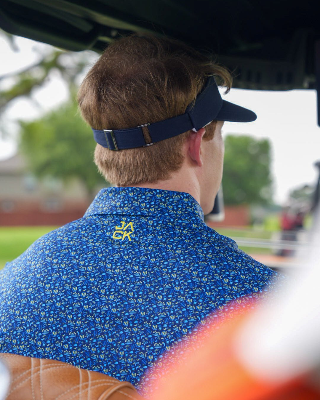 A man seen from behind wearing a blue short-sleeve button-up shirt with a yellow floral pattern and a dark navy visor, sitting in a golf cart outdoors.