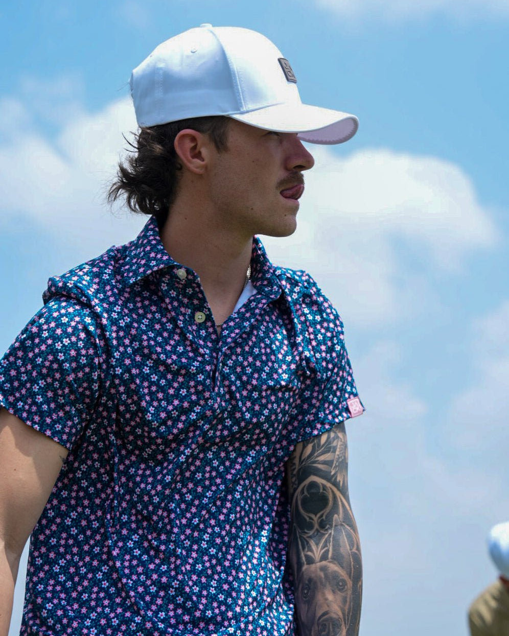 A man wearing a navy blue short-sleeve button-up shirt with a pink and white floral pattern, a white baseball cap, and a visible tattoo on his left arm, set against a bright blue sky with clouds.