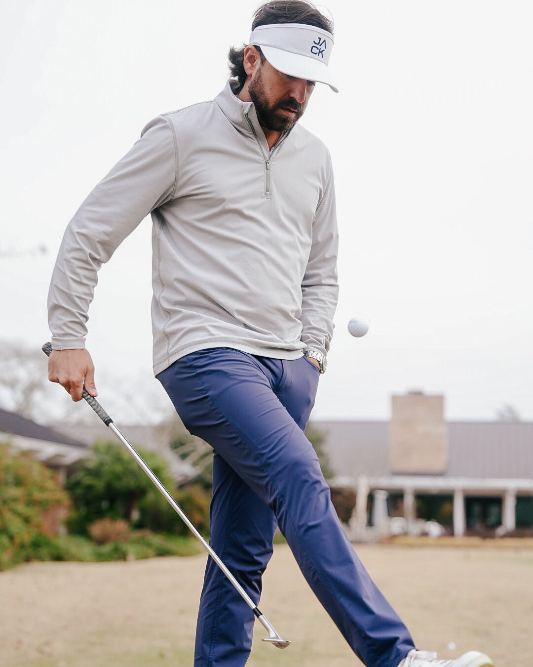 A man on a golf course wearing a light gray quarter-zip pullover, navy blue pants, and a white visor, mid-swing with a golf club.
