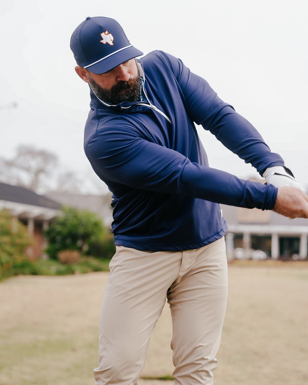 A bearded man in a navy blue long-sleeve athletic quarter-zip pullover and matching navy cap with a white logo, mid-swing on a golf course.