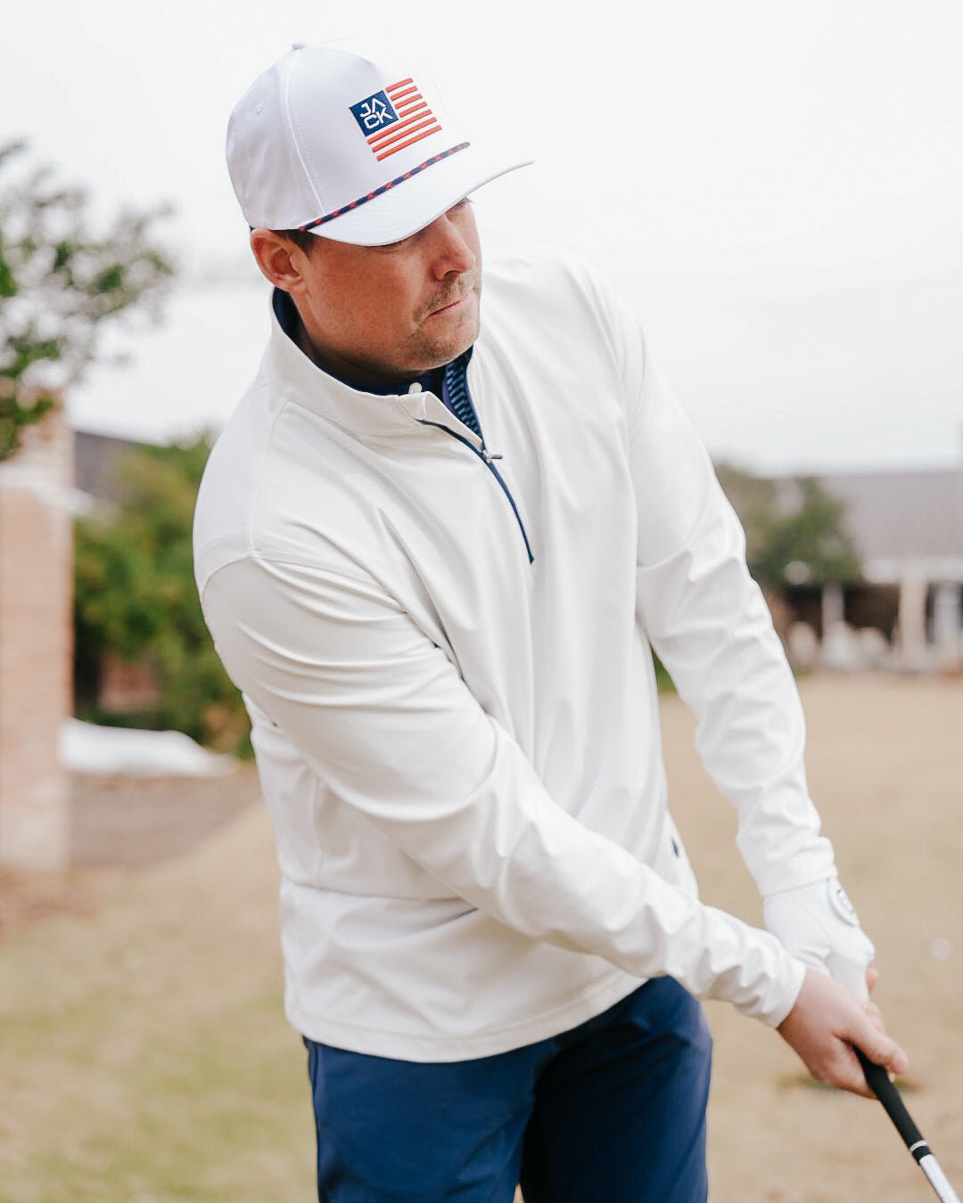 A man wearing a white long-sleeve quarter-zip pullover, a white cap with an American flag patch, and navy pants, captured mid-swing on a golf course.