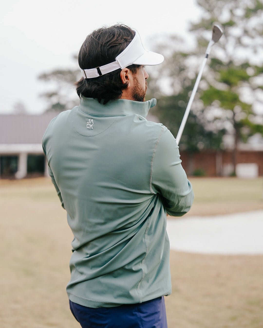 A man on a golf course, seen from behind, wearing a sage green long-sleeve performance quarter-zip pullover and a white visor while holding a golf club.
