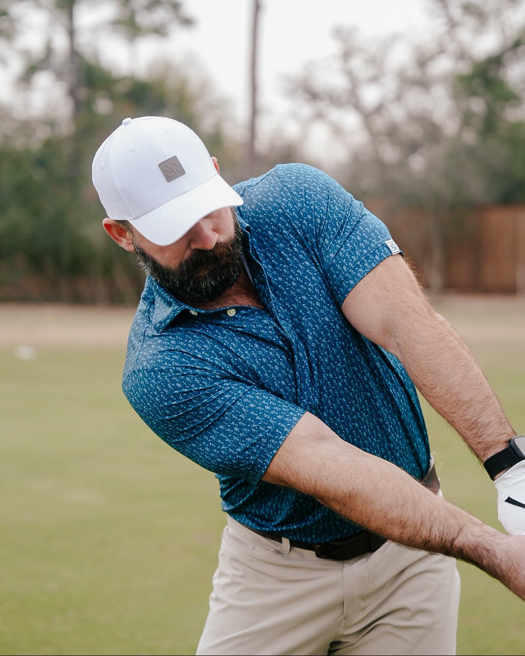 A bearded man in a blue patterned polo shirt, beige pants, and a white cap swings a golf club on a grassy course.
