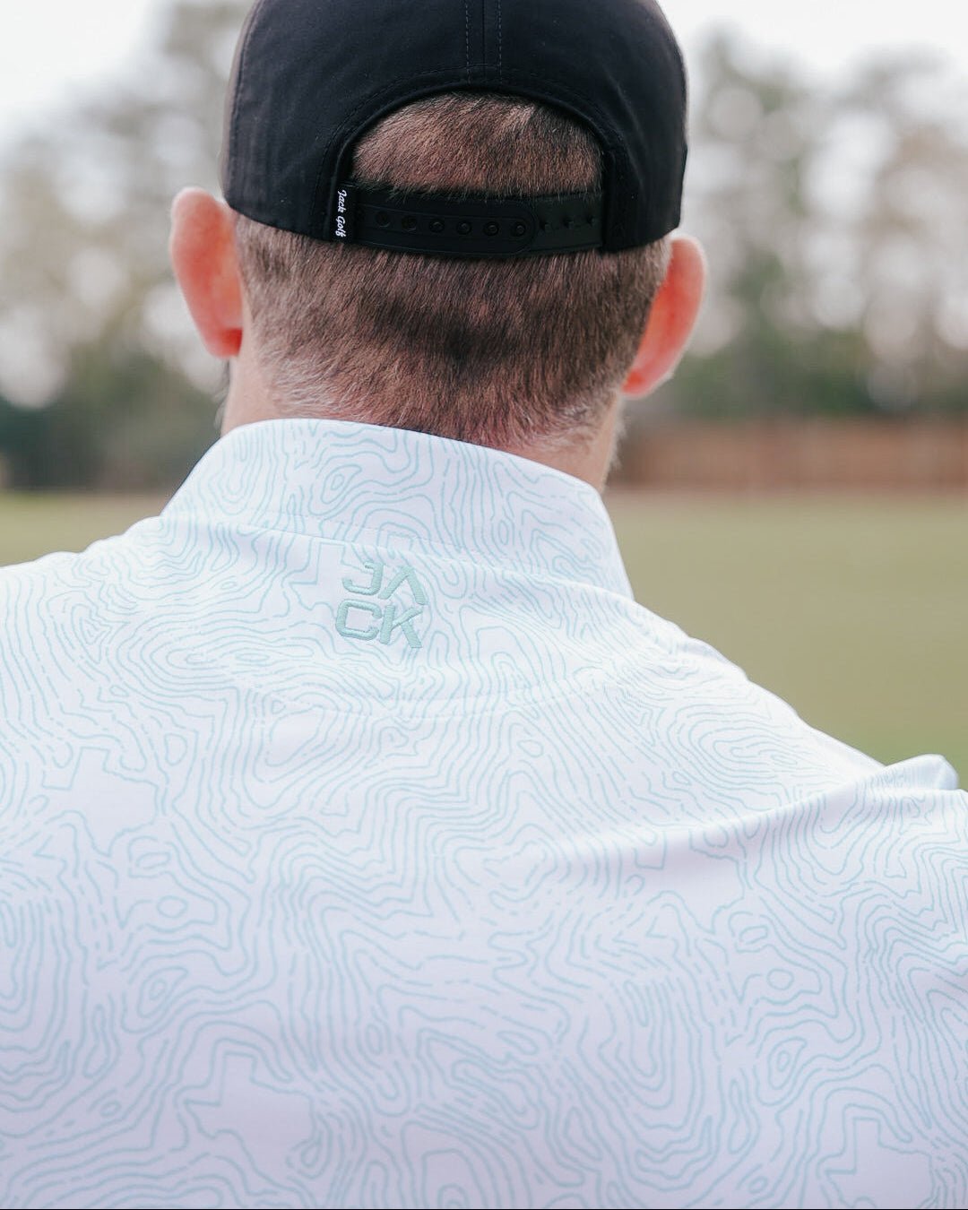 Person wearing a light blue patterned shirt with a logo on the back, standing outdoors.