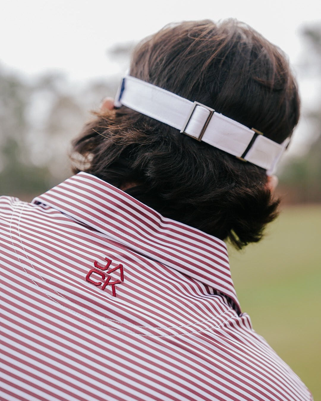 A close-up back view of a person wearing a white headband with black trim and a red-and-white striped collared shirt with a red embroidered logo.