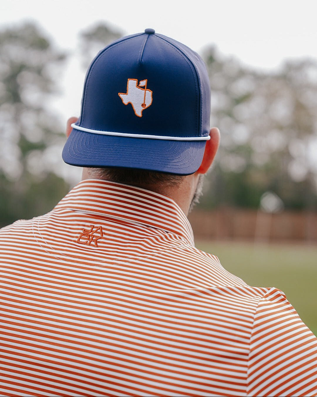 A man viewed from behind wearing a navy blue baseball cap with an orange and white Texas outline logo, paired with an orange and white striped polo shirt in an outdoor setting.