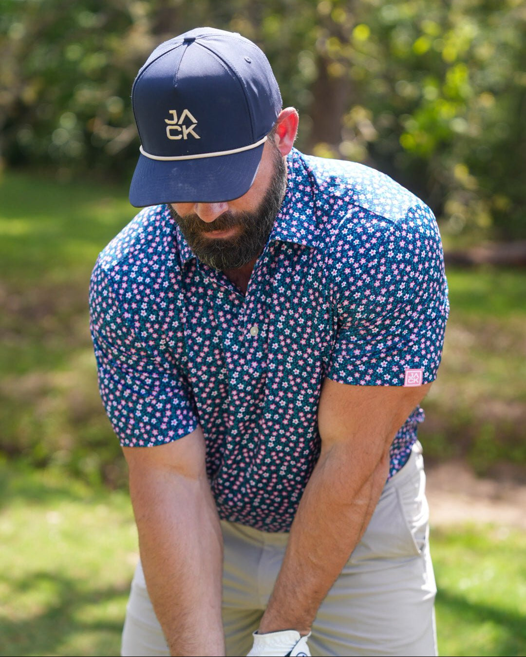 A man on a golf course wearing a navy blue short-sleeve button-up shirt with a pink and white floral pattern, a navy blue cap with a gold logo, and light khaki pants.
