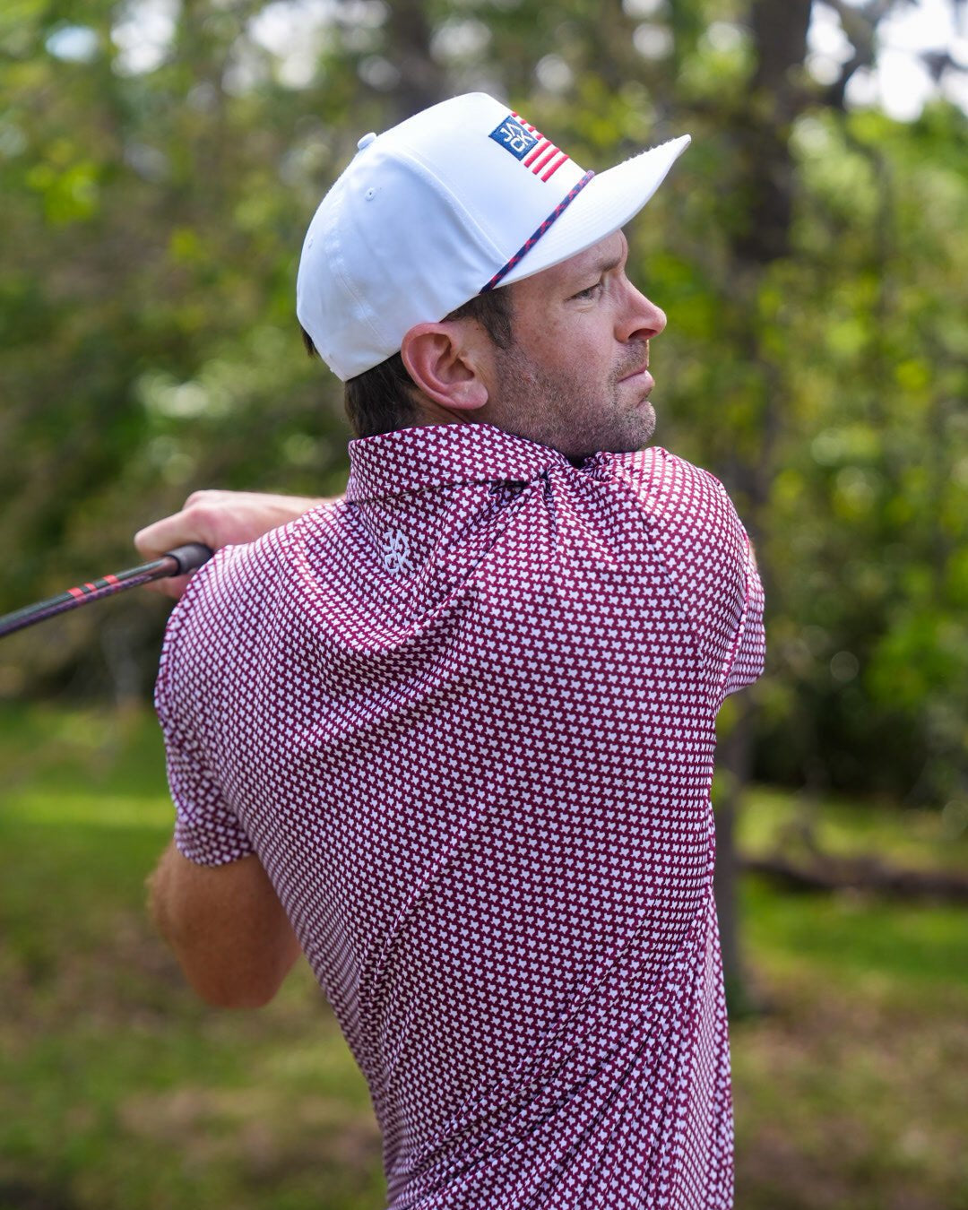 A man wearing a white baseball cap with an American flag patch and a pink-and-white patterned polo shirt, captured mid-swing on a golf course with green trees in the background.