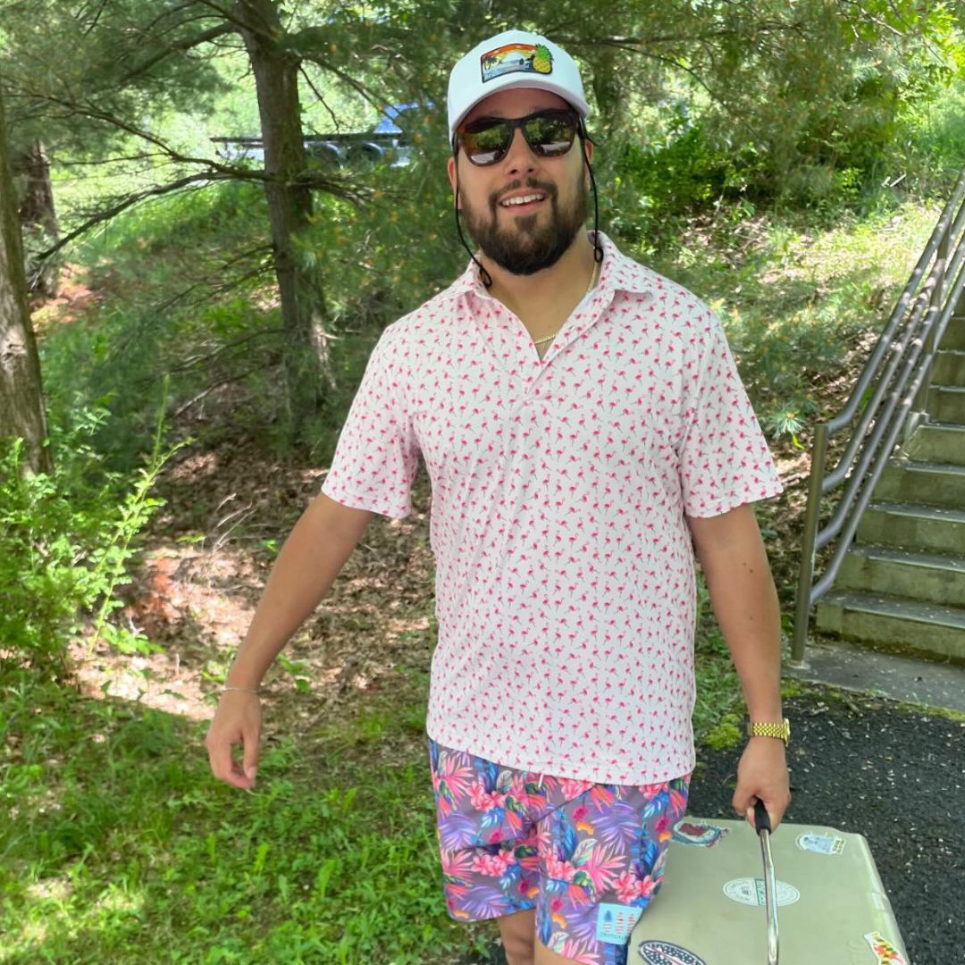 Man in a pink patterned shirt and colorful shorts holding a suitcase outdoors.