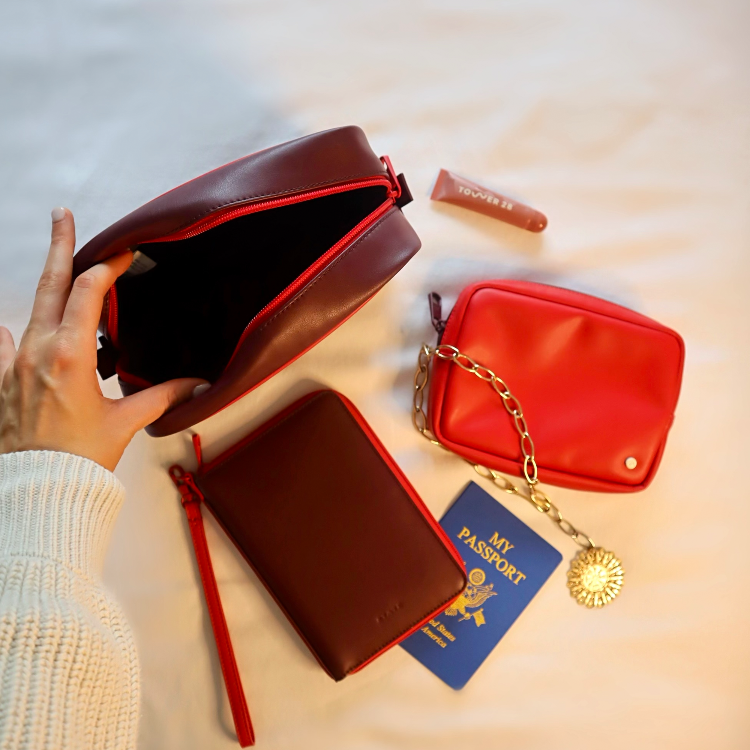 A hand holds open a dark brown leather cosmetic pouch with red trim, placed on a light fabric surface alongside a red zip pouch with a gold chain, a brown zip case with a wrist strap, a blue passport, and a lip product tube.