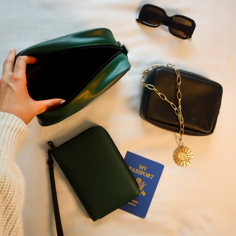 A styled flat-lay of two dark green and black faux-leather accessories on a white fabric background, including a hand holding open the green pouch, a black pouch with a gold chain and sun pendant, a green wristlet wallet, and a blue passport. A pair of black sunglasses is also present.