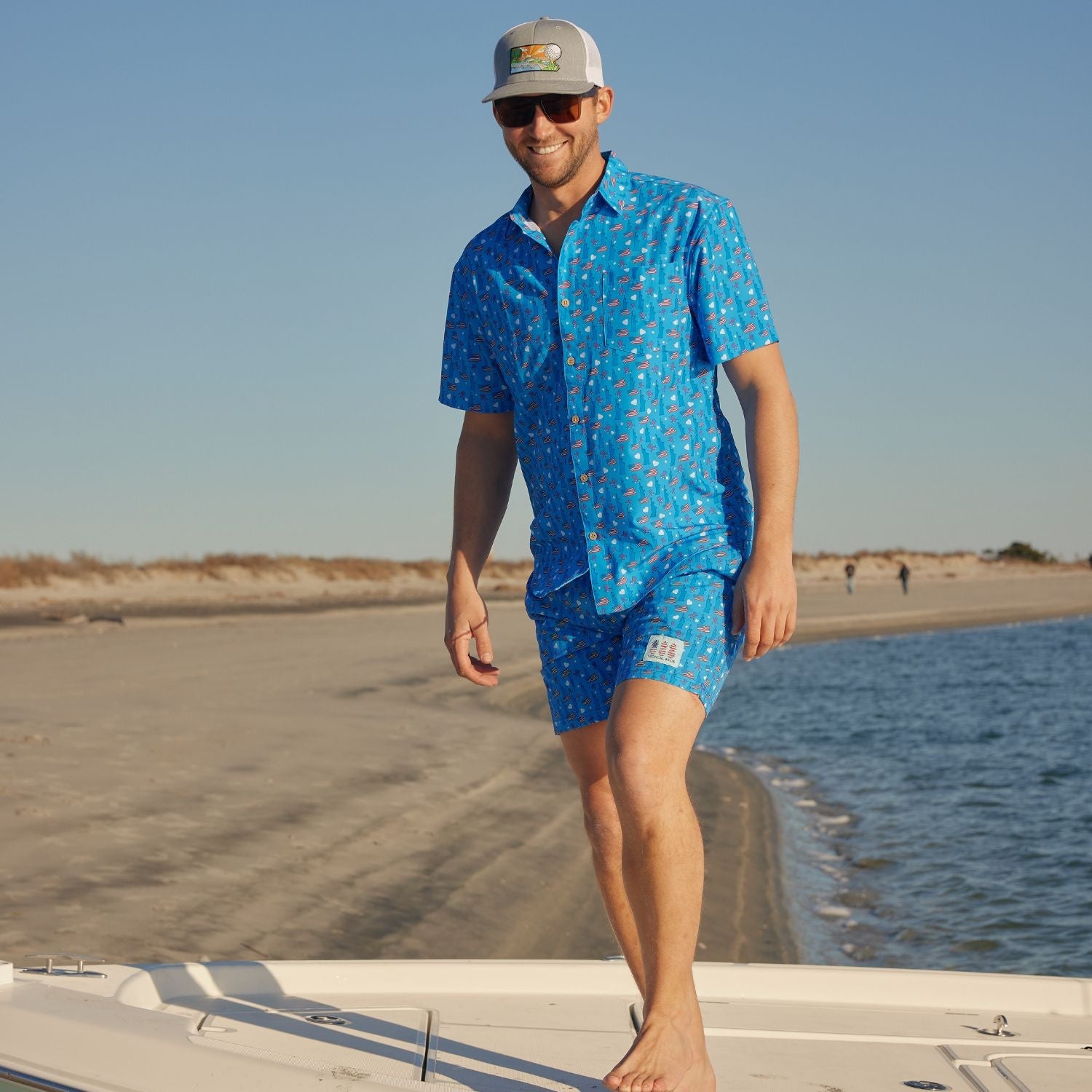 Man in blue patterned shirt and shorts standing on a boat at the beach.