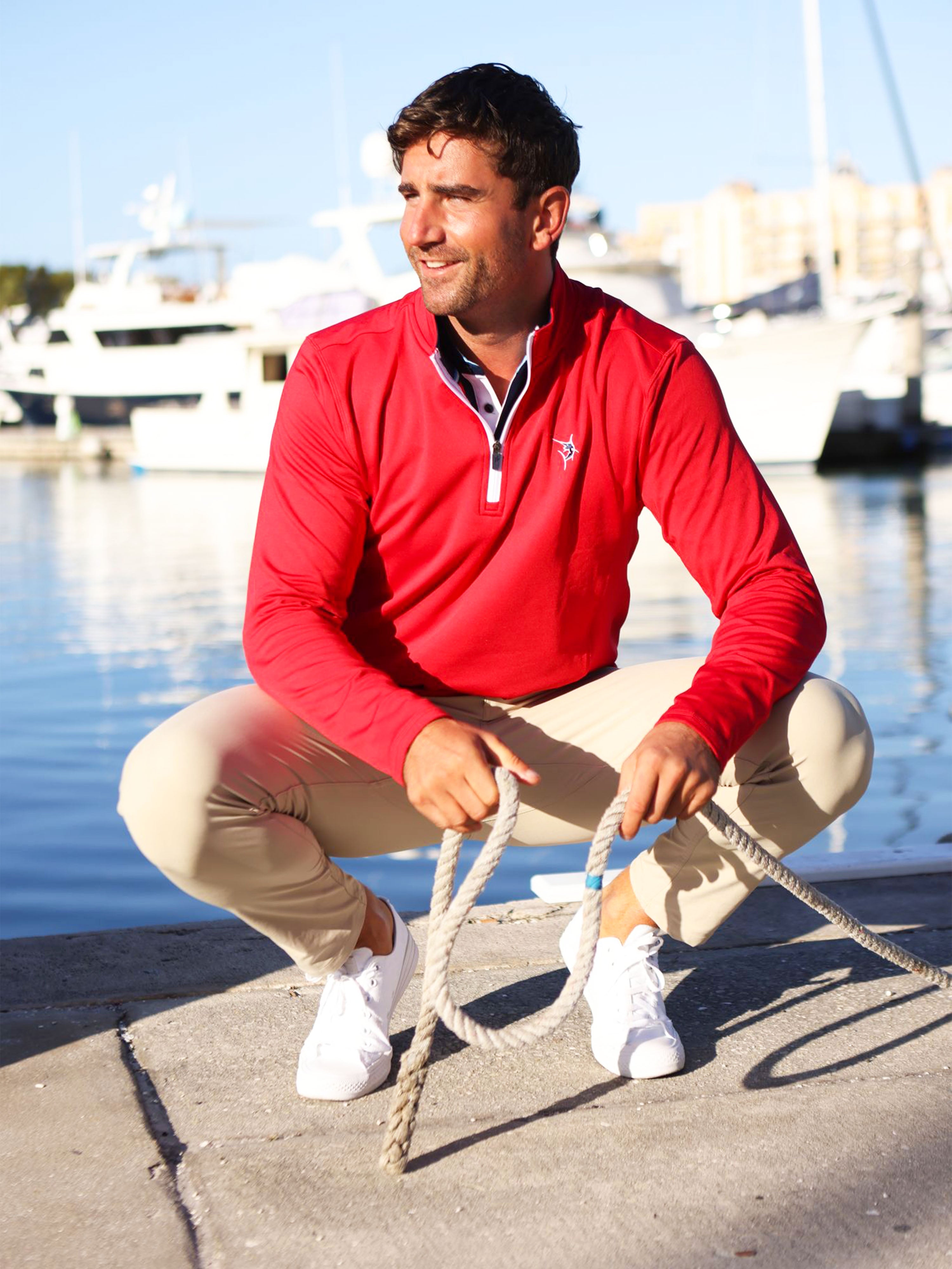 Man in a red shirt sitting on a dock holding a rope, with boats in the background.