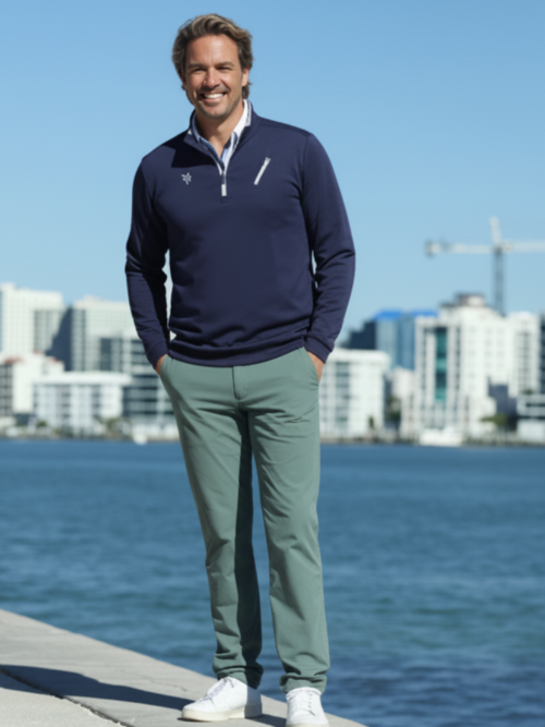 Man wearing a navy sweater and green pants standing by a waterfront with buildings in the background