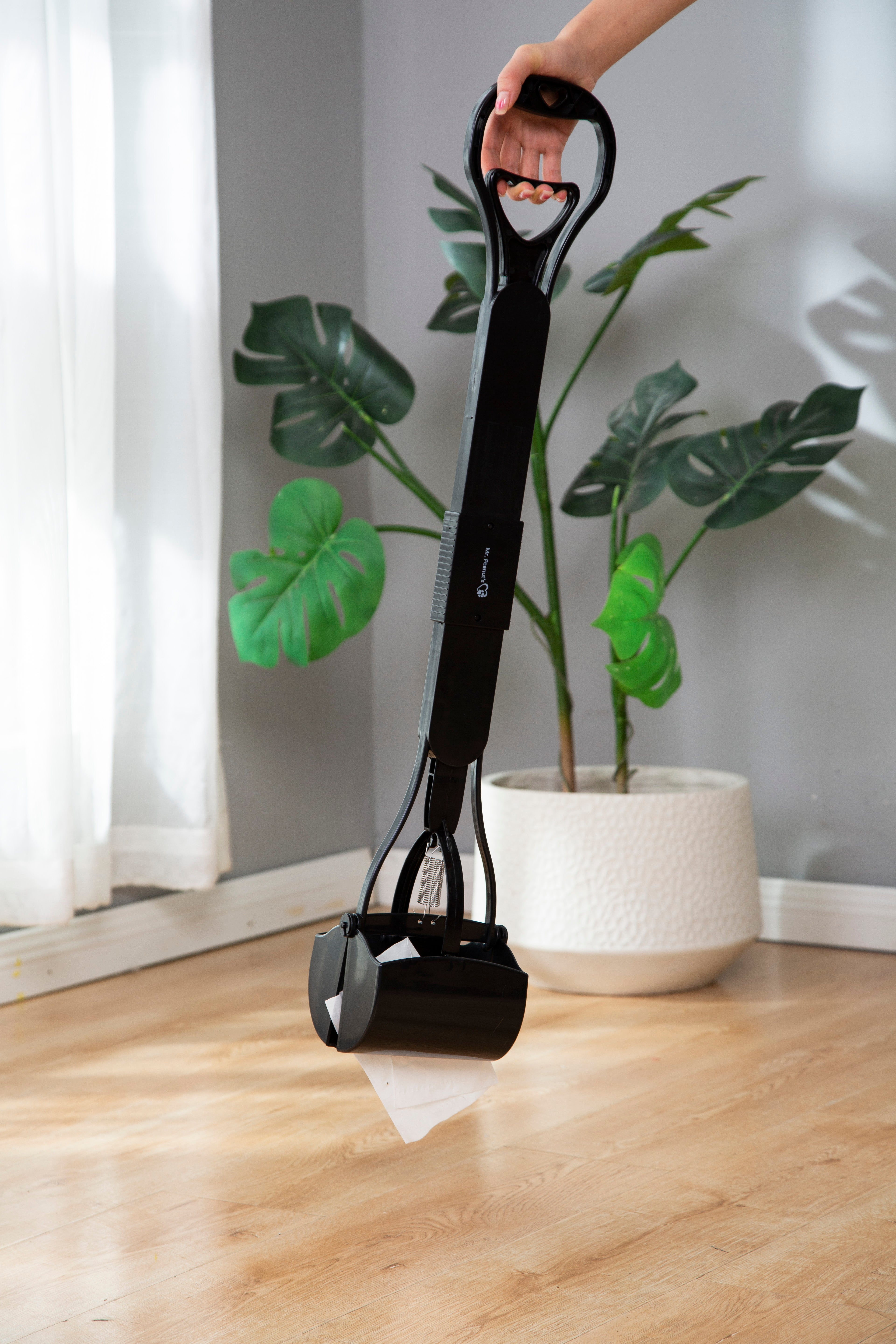 Black pet waste bag dispenser being used on a wooden floor with a plant in the background