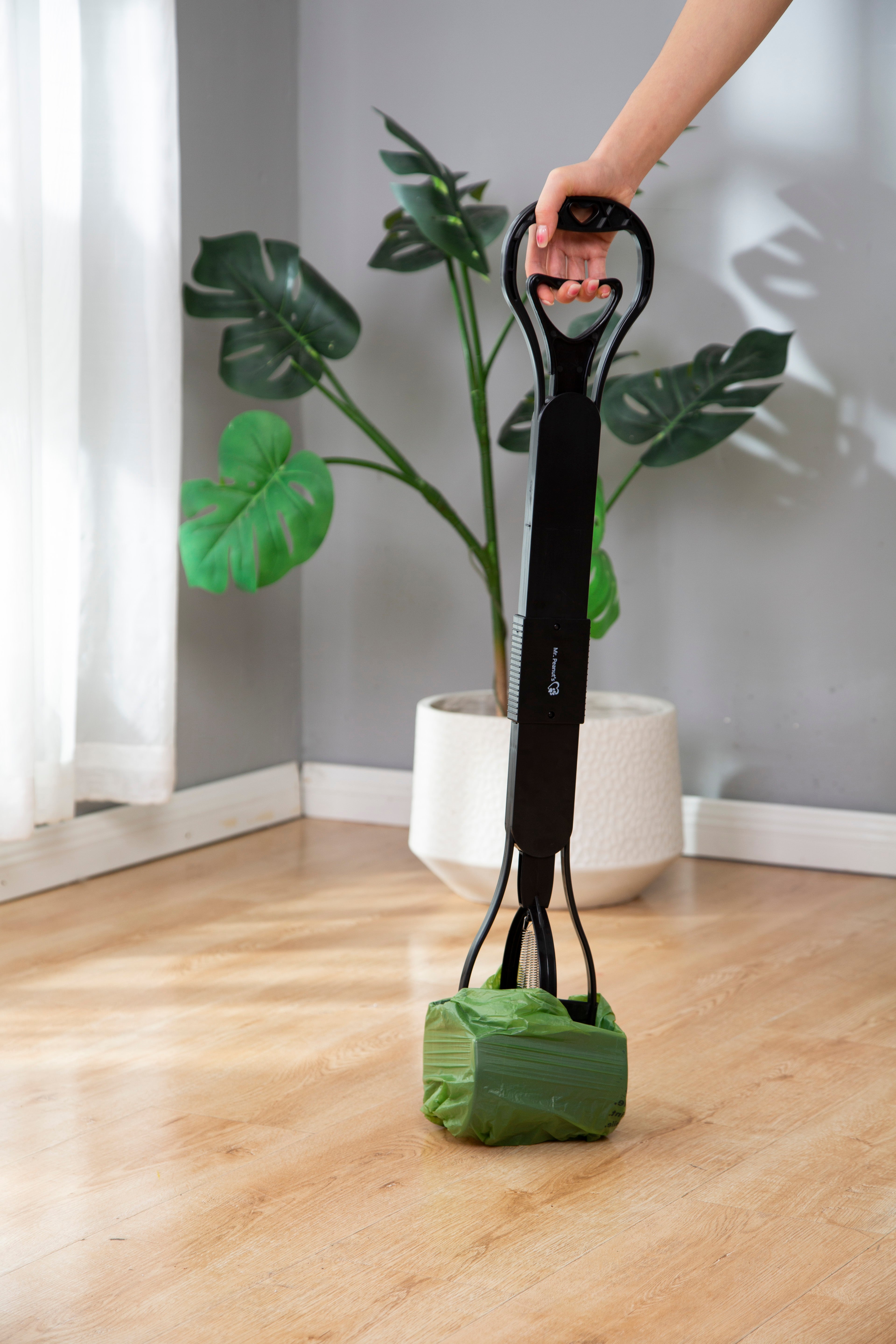 Person holding a black strap with a green bag attached, standing in a room with a plant and white curtains.