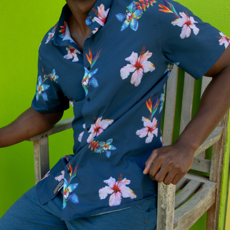 A man wearing a dark teal blue short-sleeve button-up shirt with a colorful tropical hibiscus floral print, paired with matching blue shorts, sitting on a rustic wooden chair against a bright lime green background.