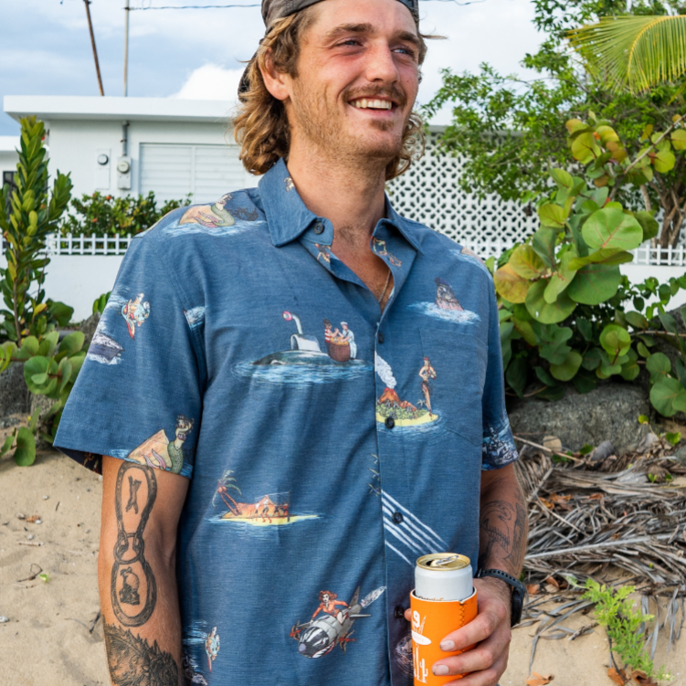 Man wearing a blue patterned shirt holding an orange can on a beach.