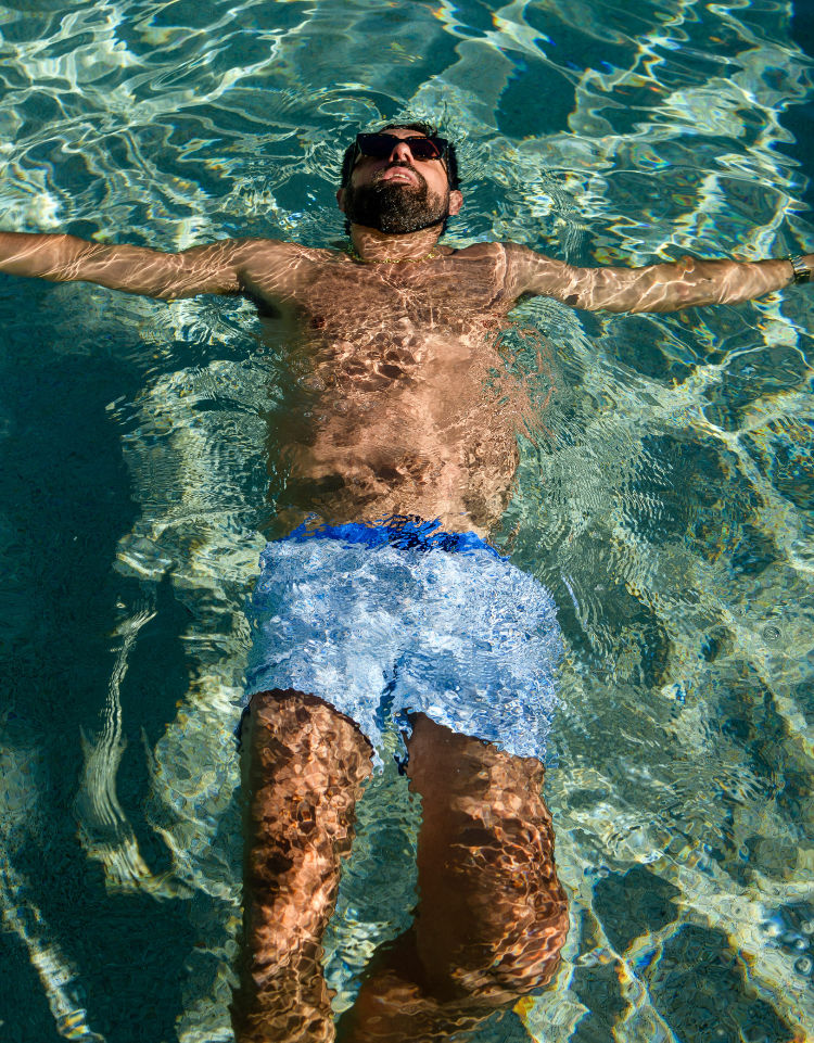 A man floating on his back in a swimming pool, wearing light blue swim trunks with a darker blue waistband and dark sunglasses. The water is clear and reflects sunlight, showing the pool floor underneath.