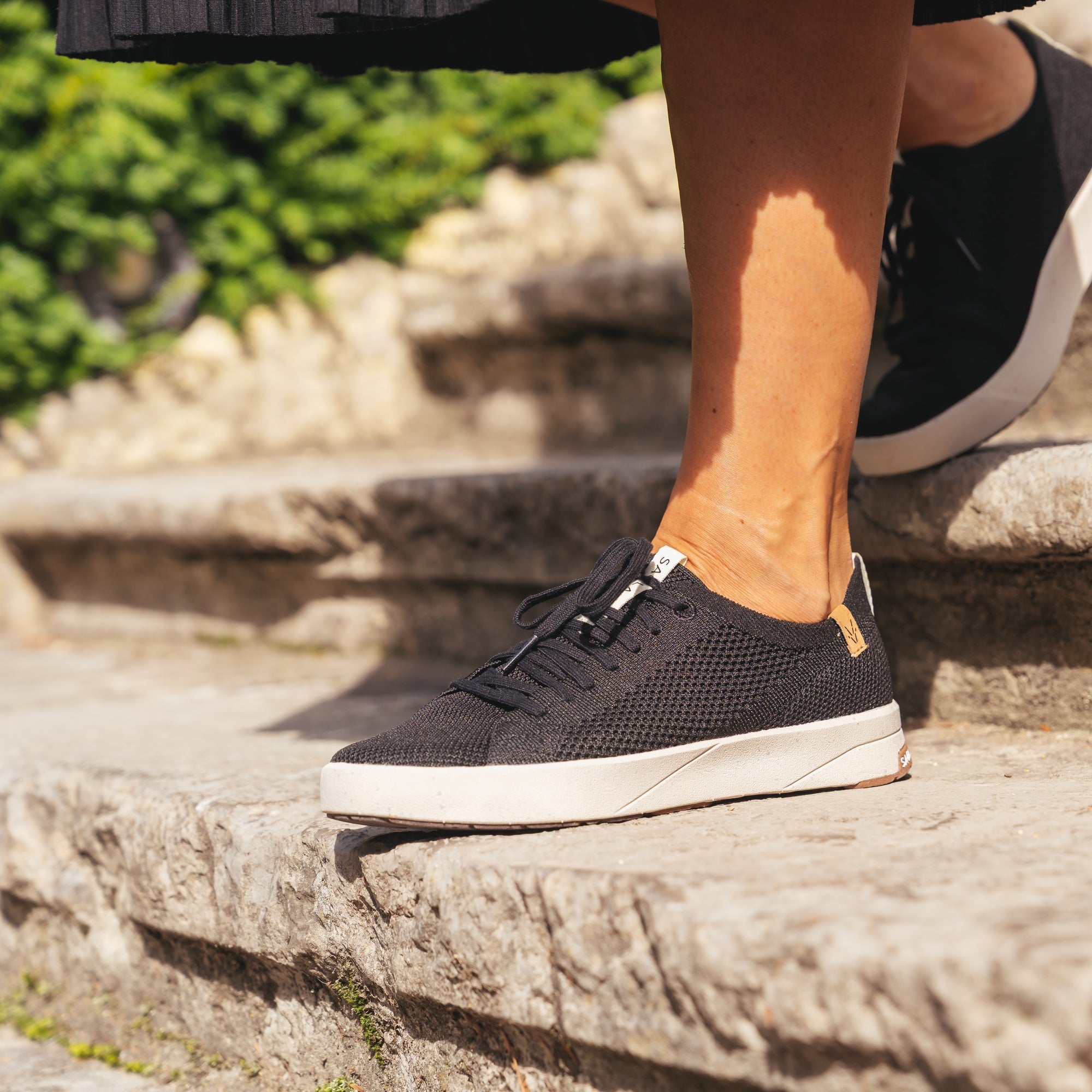 A person wearing black knit slip-on sneakers with white soles, standing on stone steps outdoors.
