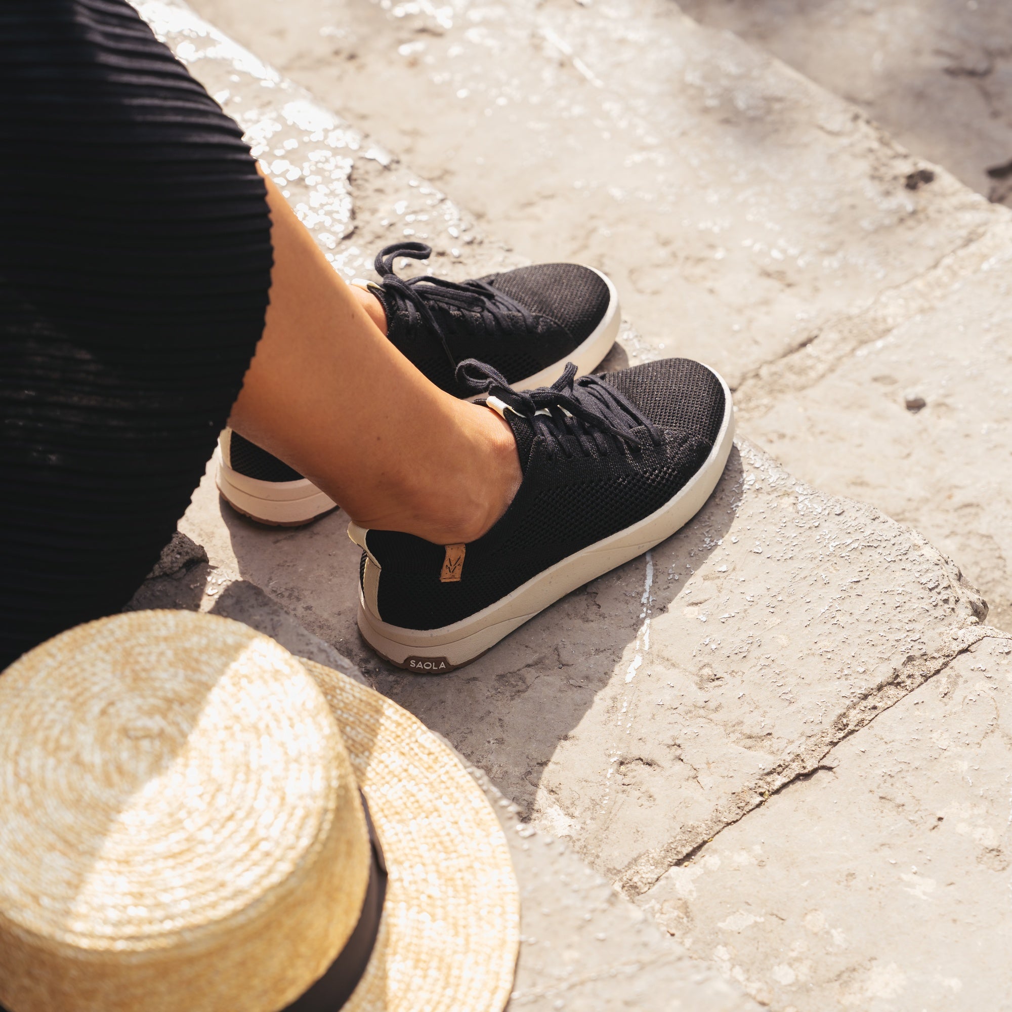 A person wearing black knit sneakers with off-white soles sits on stone steps, with a straw hat partially visible in the foreground. The shoes show the Fila brand name on the heel.