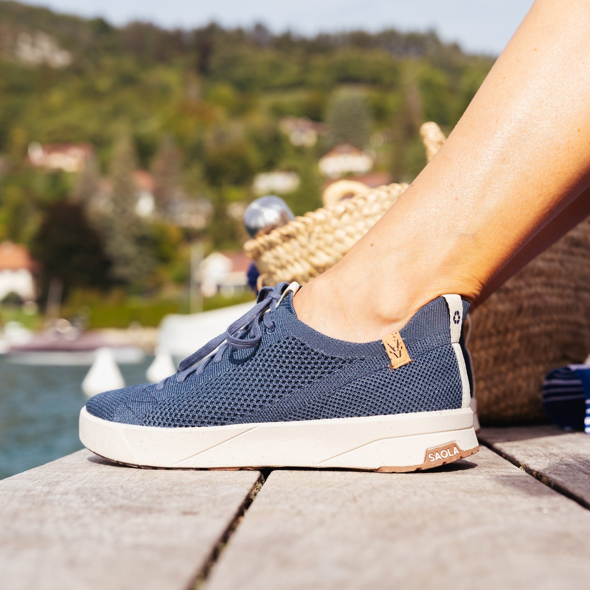 A person's foot wearing a navy blue knit slip-on shoe with a white sole, resting on a wooden dock with a scenic lake and green hills in the background.