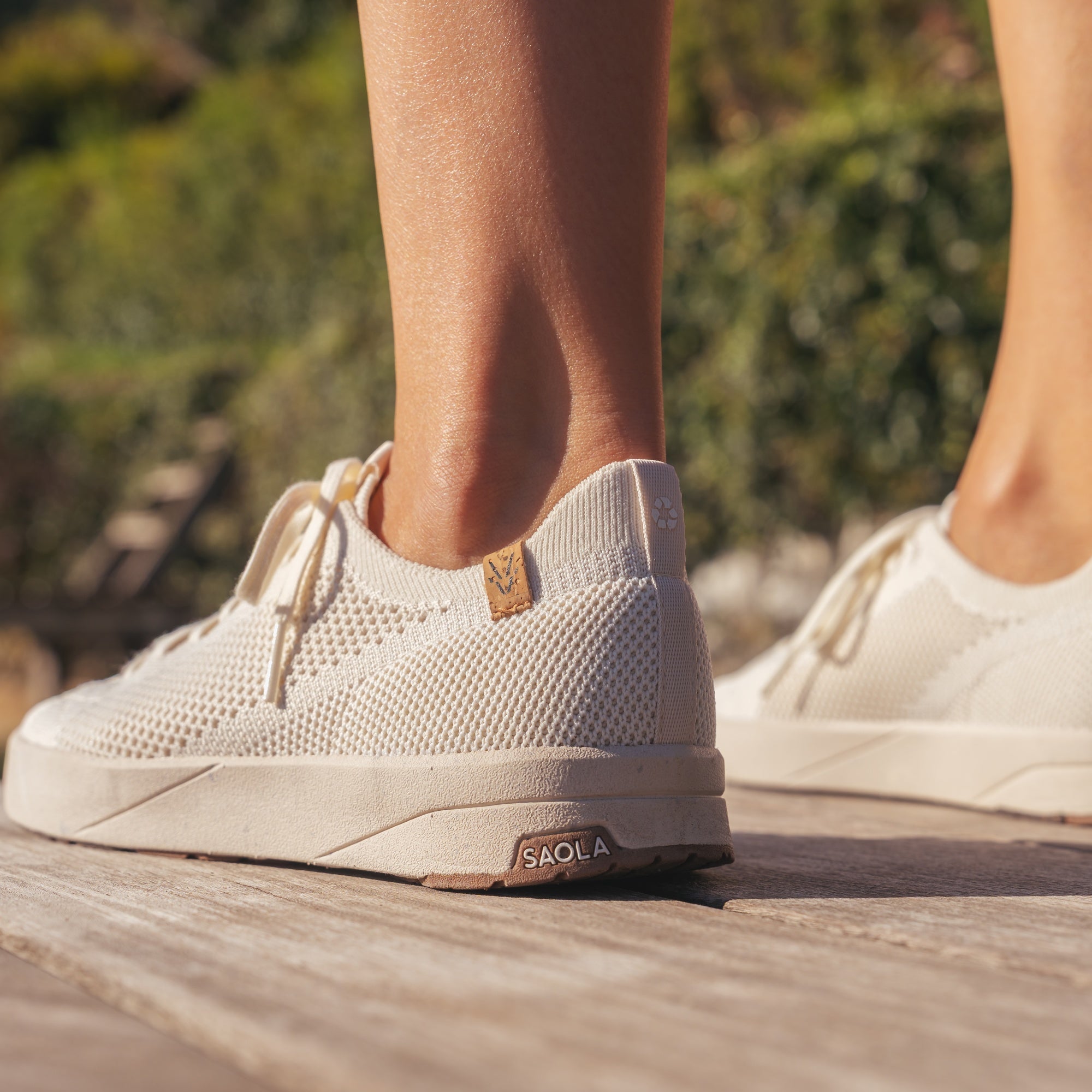 A person wearing off-white knit sneakers with beige laces and a small tan brand tab, standing on a wooden deck outdoors. The brand name SAOLA is visible on the heel of the shoe.