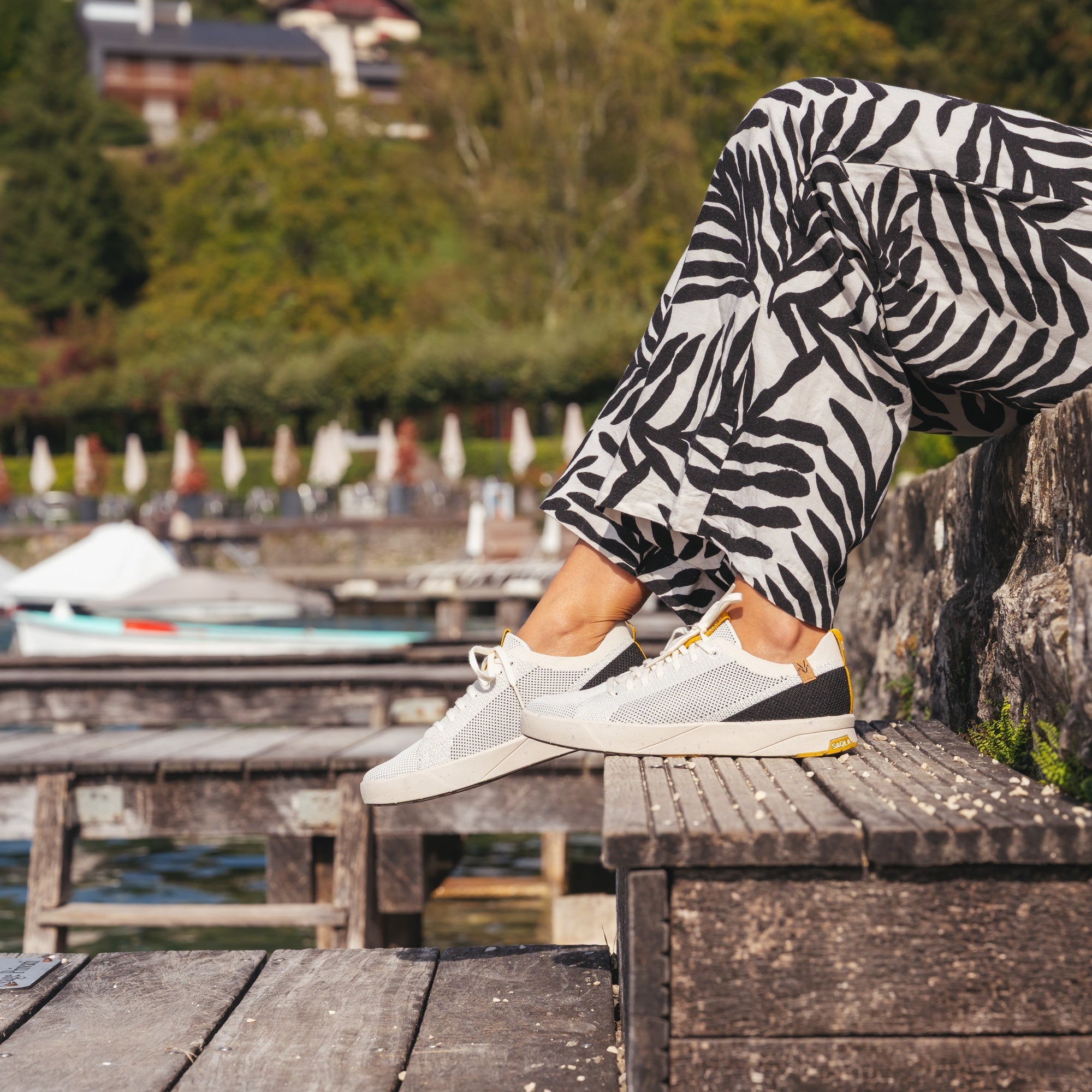 A person sitting on a wooden dock by a lake, wearing white sneakers with black and gray panels and black-and-white patterned pants. The background shows a blurred outdoor harbor scene with boats and trees.
