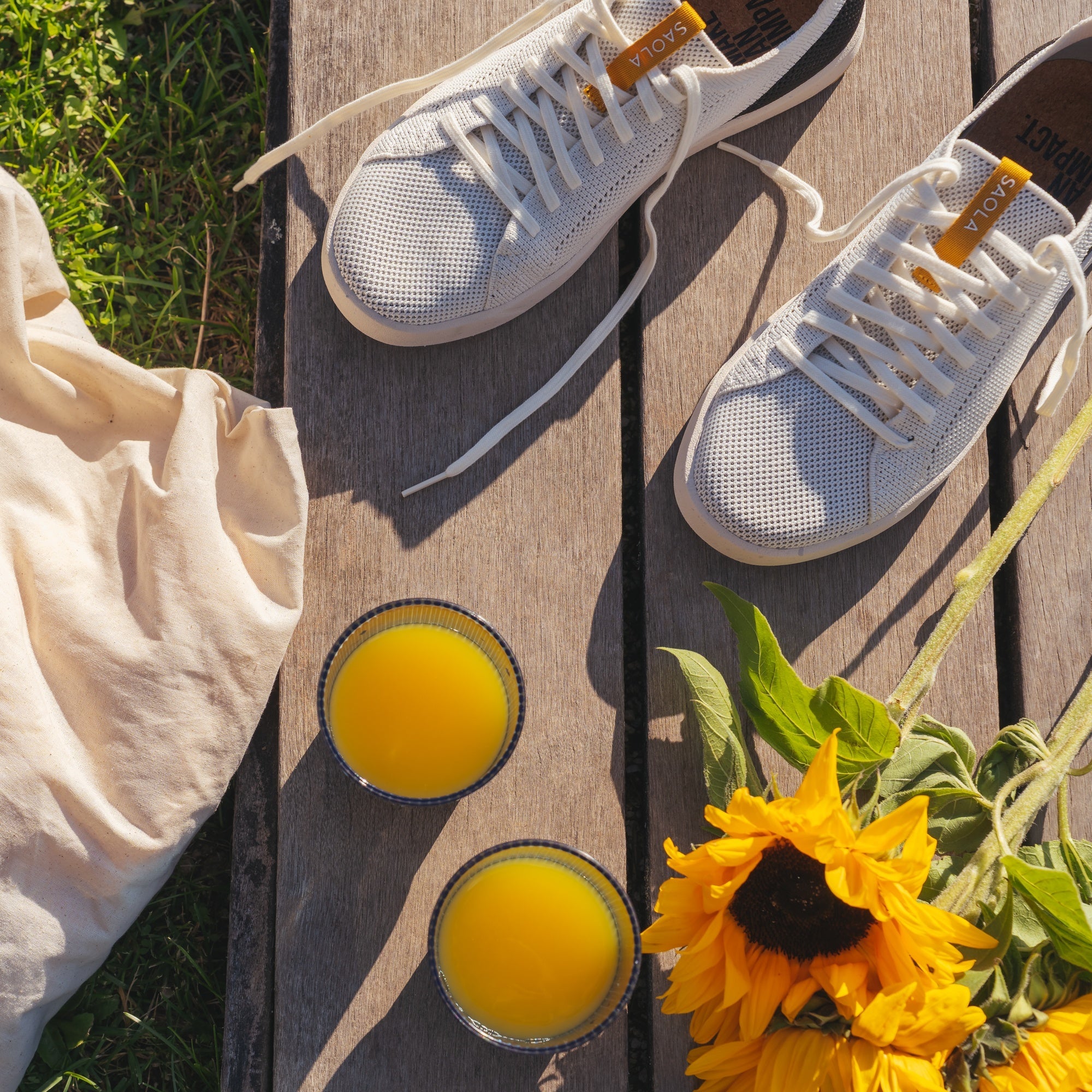 A pair of white low-top sneakers with white laces and small orange accents, arranged on a wooden deck outdoors with two glasses of orange juice, a cream fabric bag, and yellow sunflowers nearby.