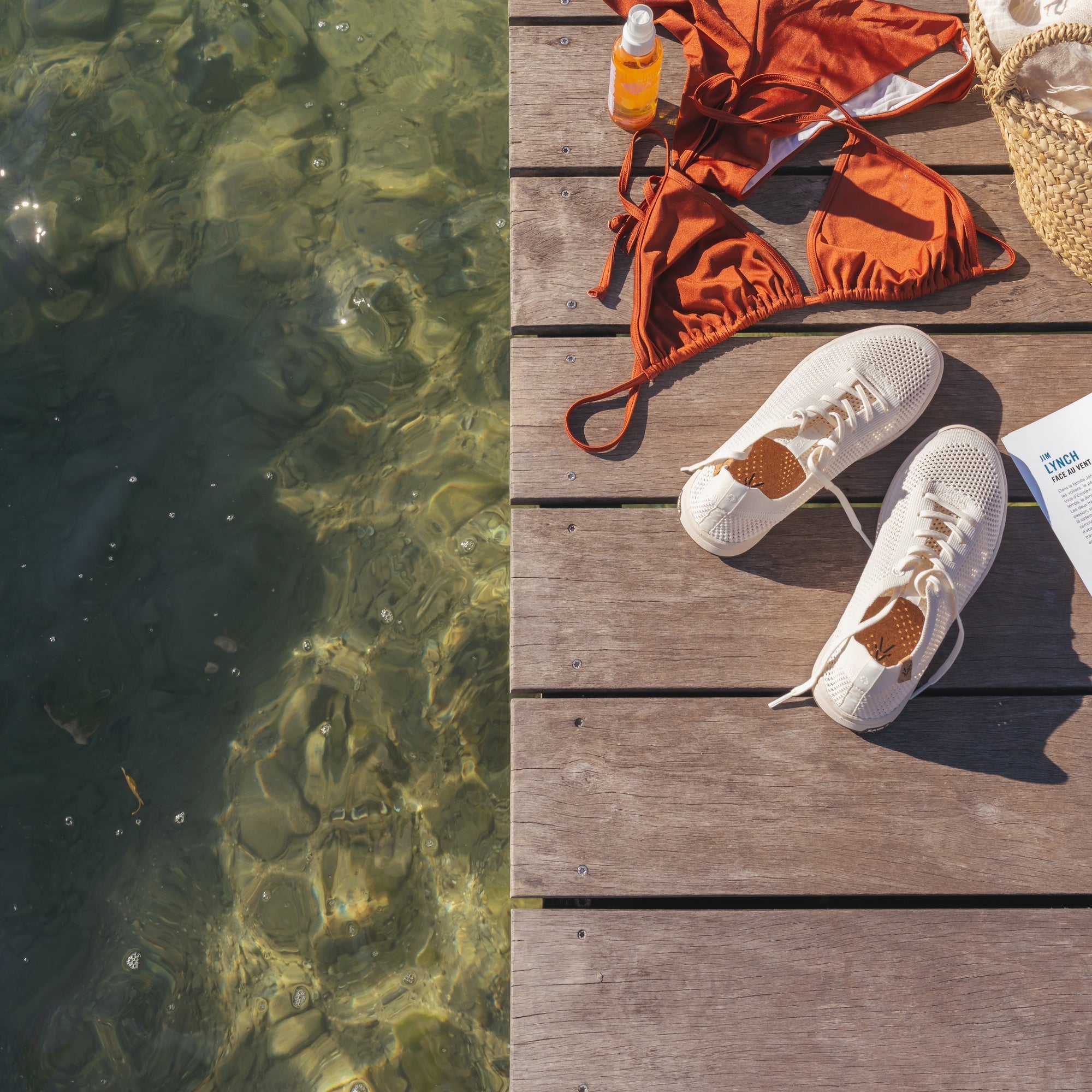 A styled beachside flat-lay on a wooden dock next to clear water, featuring an orange bikini, white sneakers, a straw hat, a bottle of sunscreen, and a magazine.