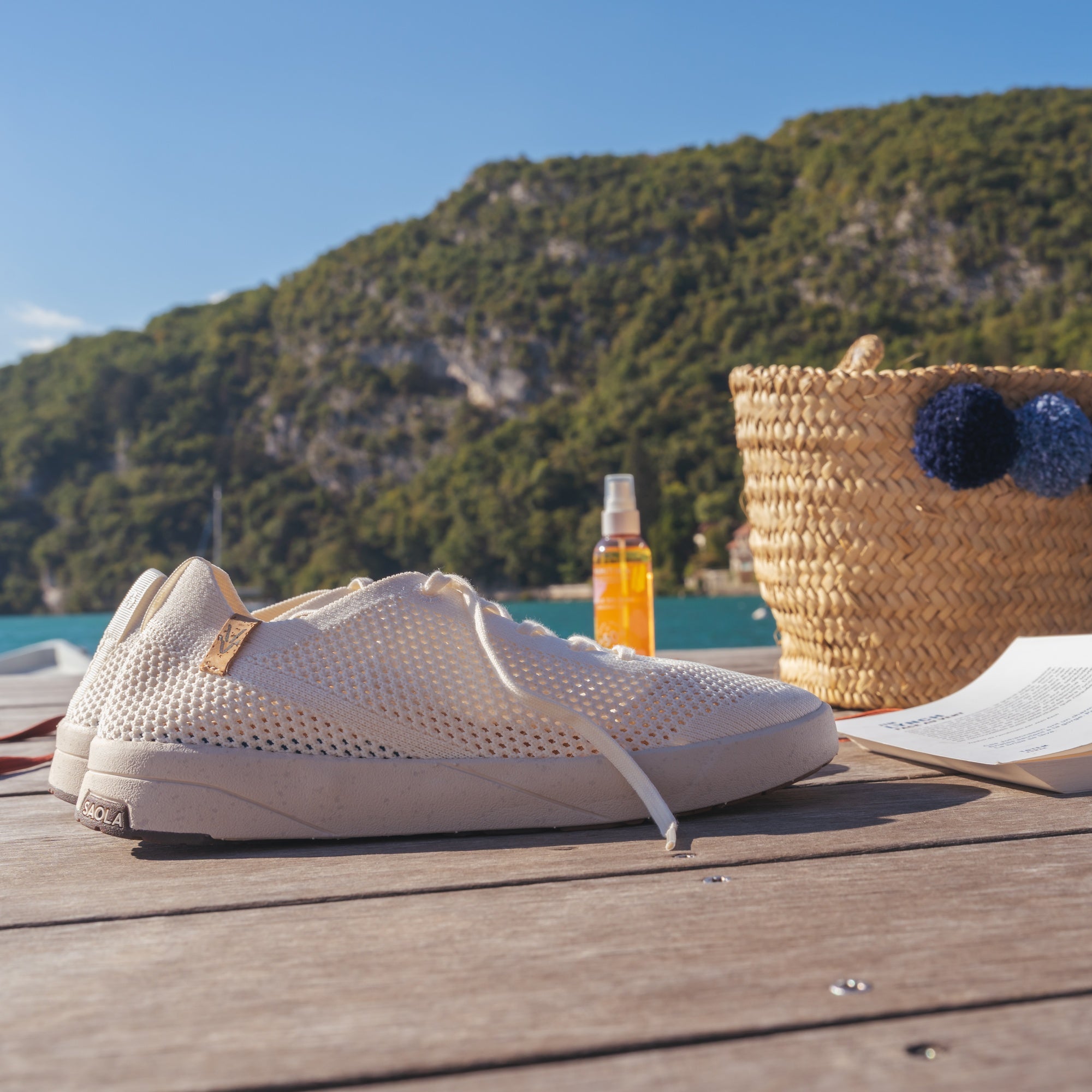 A pair of off-white mesh sneakers resting on a wooden deck with a scenic background of a green mountain, blue sea, and a straw tote bag.