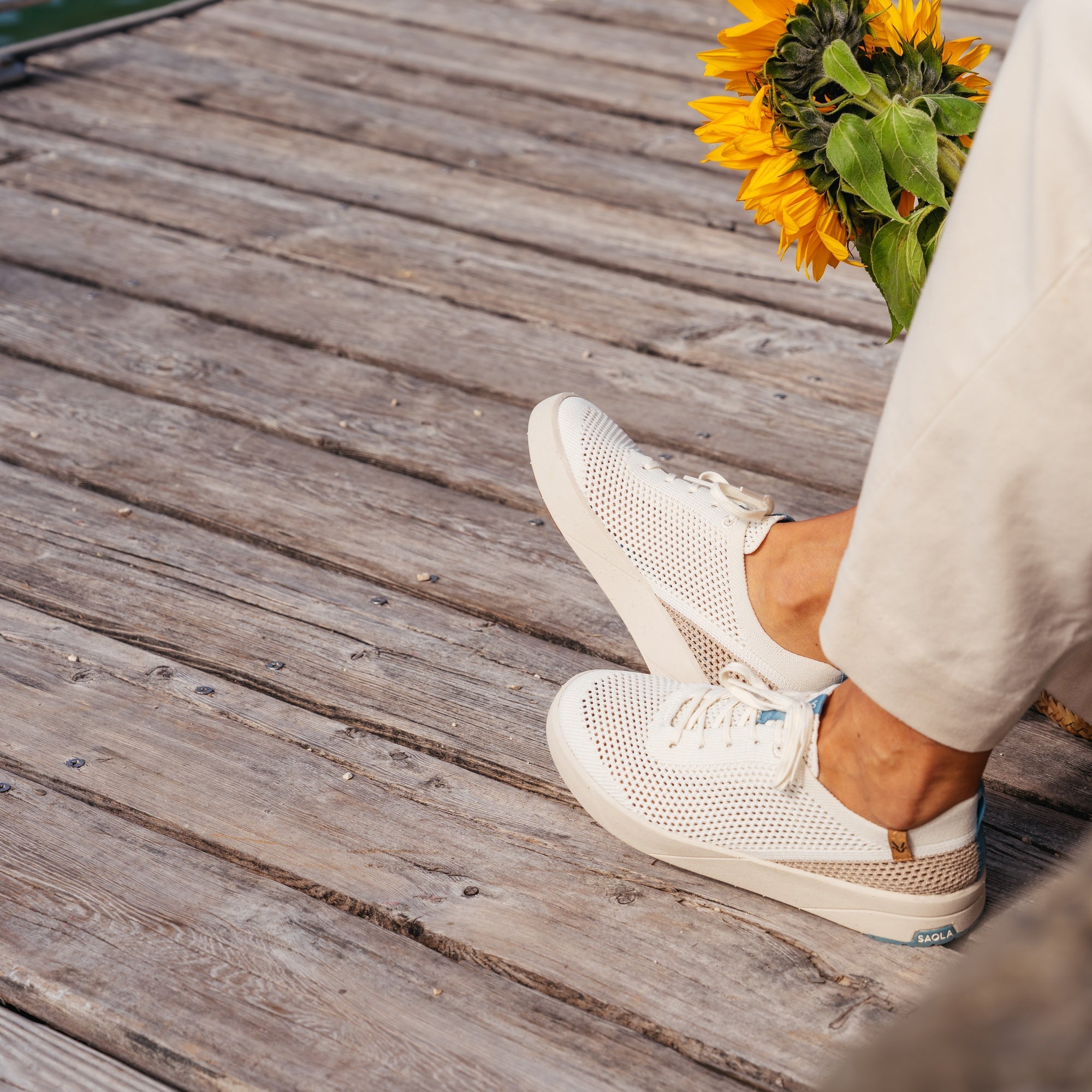 A person wearing white perforated slip-on sneakers with light beige pants, standing on weathered wooden planks and holding a bouquet of yellow sunflowers.