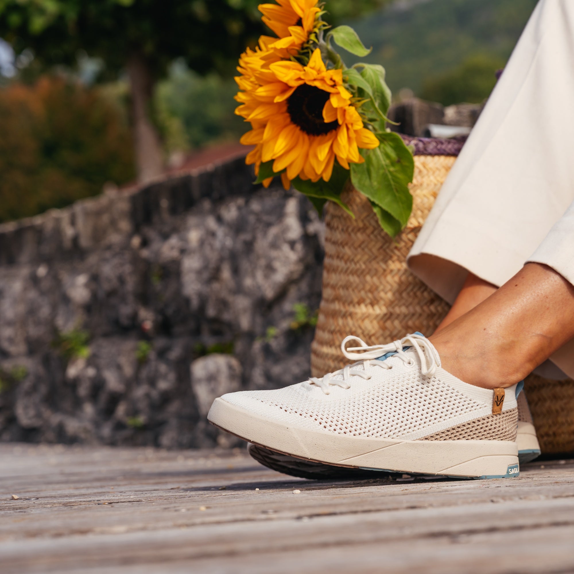 A person wearing a white perforated lace-up shoe with a beige side accent, seated outdoors near a woven basket of sunflowers on a wooden deck.