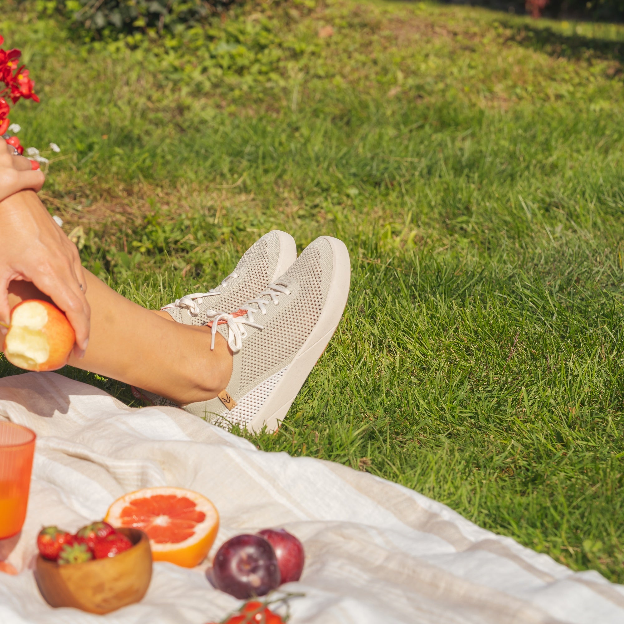 A person sitting outdoors on a white picnic blanket on green grass, wearing white mesh sneakers and holding an apple. A cup, fruit bowl, and flowers are visible in the foreground.