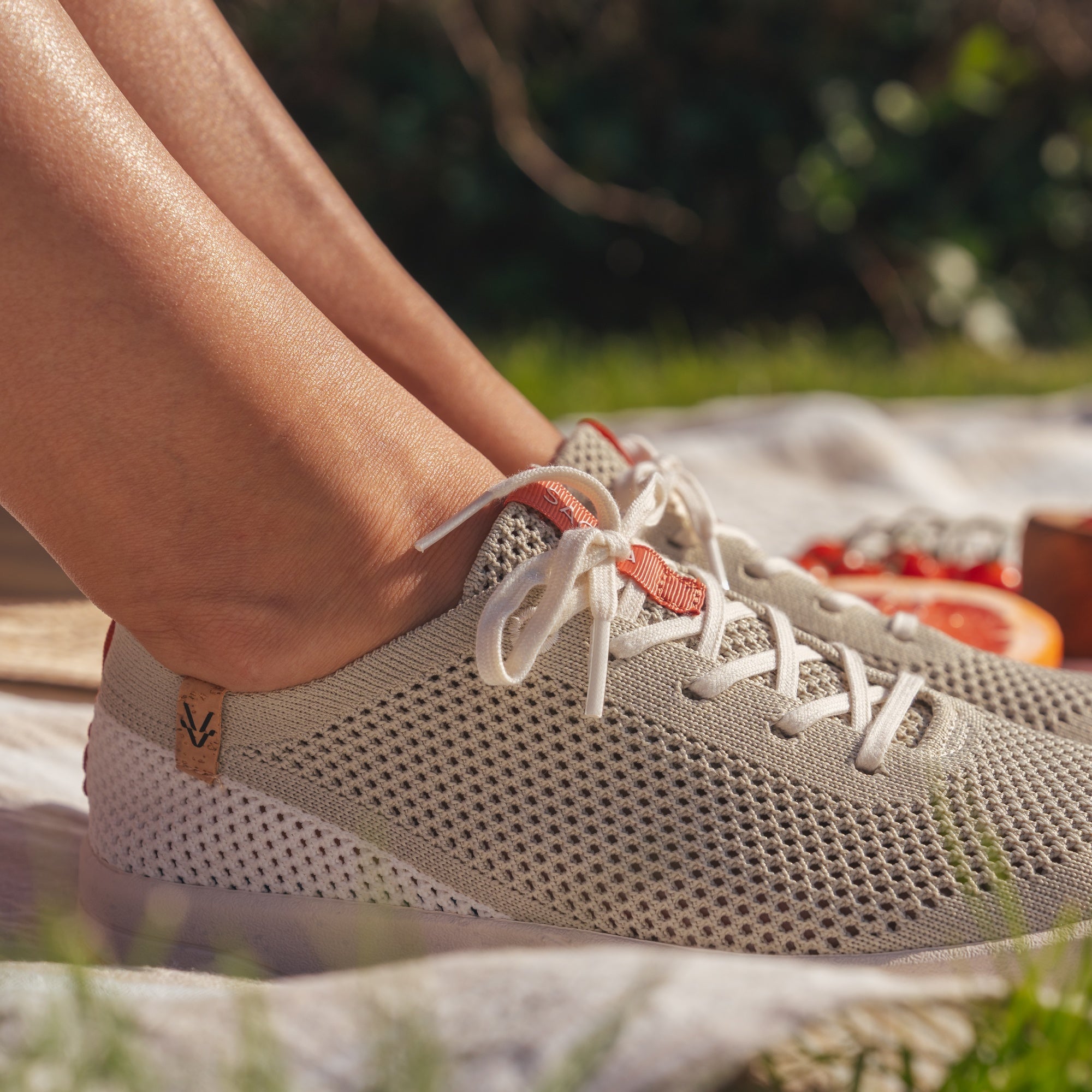 Close-up of a person's feet wearing light beige knit sneakers with white laces, sitting outdoors on a blanket with blurred greenery in the background.
