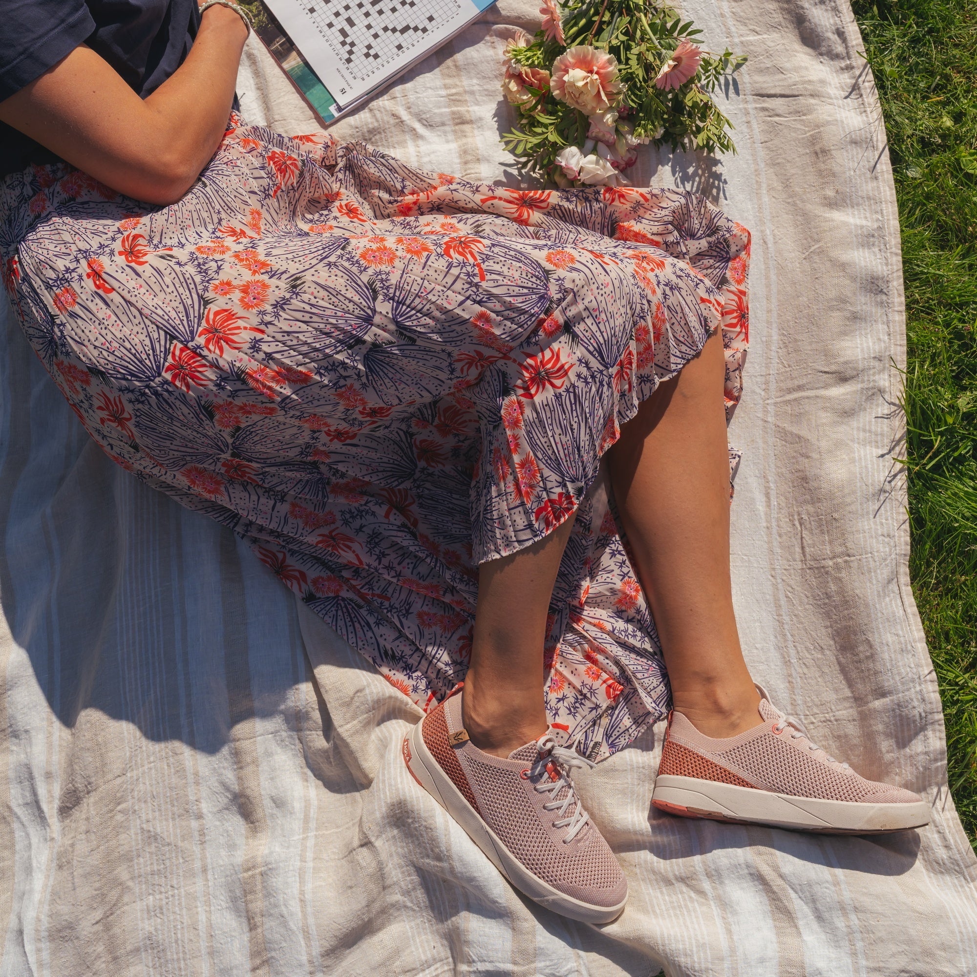 A person sitting outdoors on a white picnic blanket, wearing a loose floral midi skirt and light pink slip-on sneakers. A bouquet of flowers and a book are placed beside them on the blanket, with grass visible around the edges.