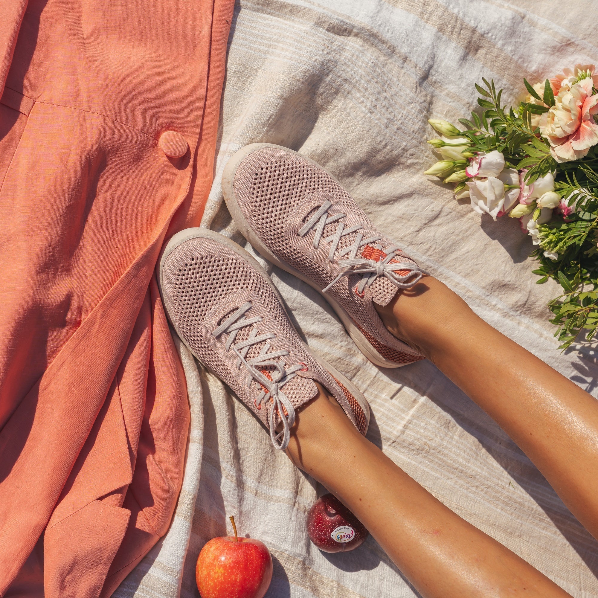 A top-down lifestyle shot of a person wearing light pink mesh sneakers with white laces, lying on a white linen blanket with a coral dress, flowers, and an apple nearby.