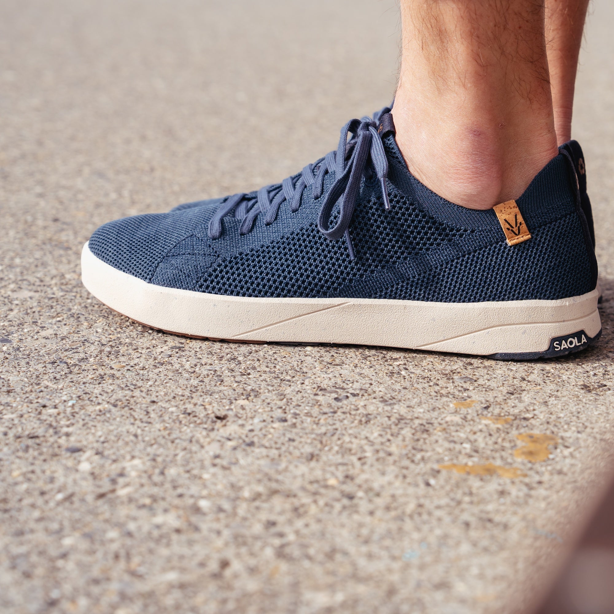 A close-up of a person's foot wearing a dark blue knit sneaker with a cream-colored midsole, standing on a textured concrete surface. The brand name 'SAOLA' is visible on the heel.