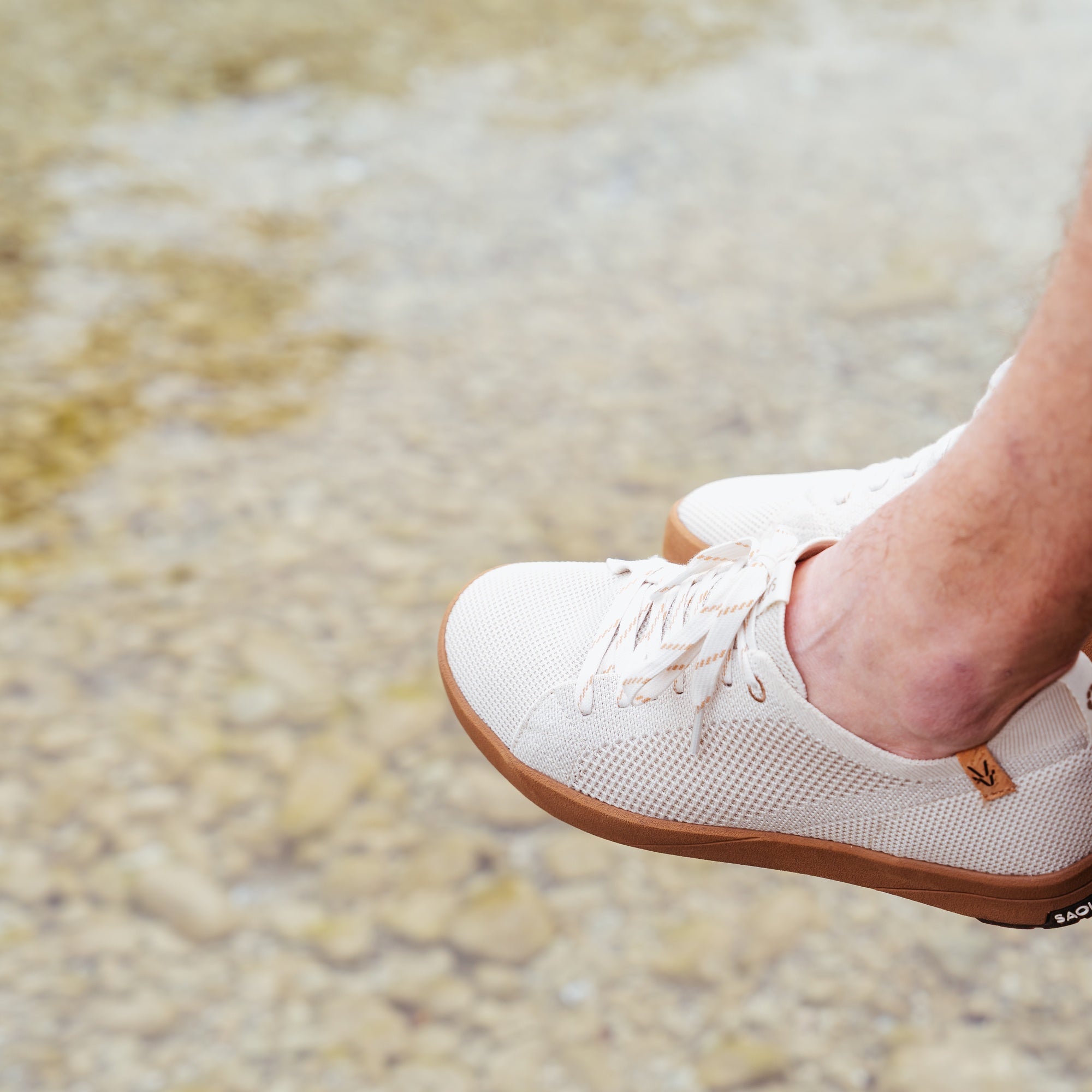 White mesh low-top sneakers with gum soles, shown on a person's feet against a blurred outdoor stone background.