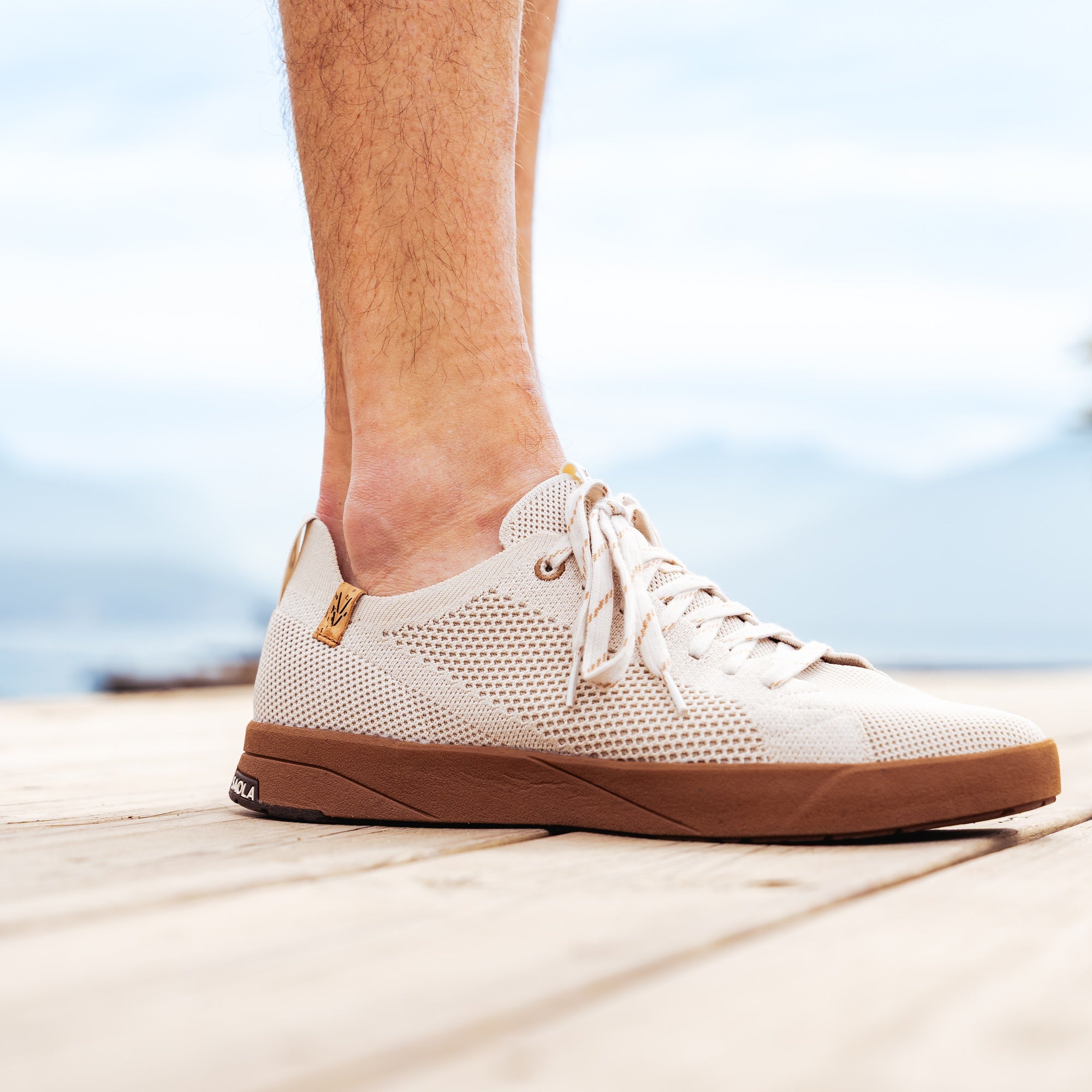 A person's foot wearing a low-top mesh sneaker with a brown gum sole, standing on a light wooden deck with a blurred outdoor background.