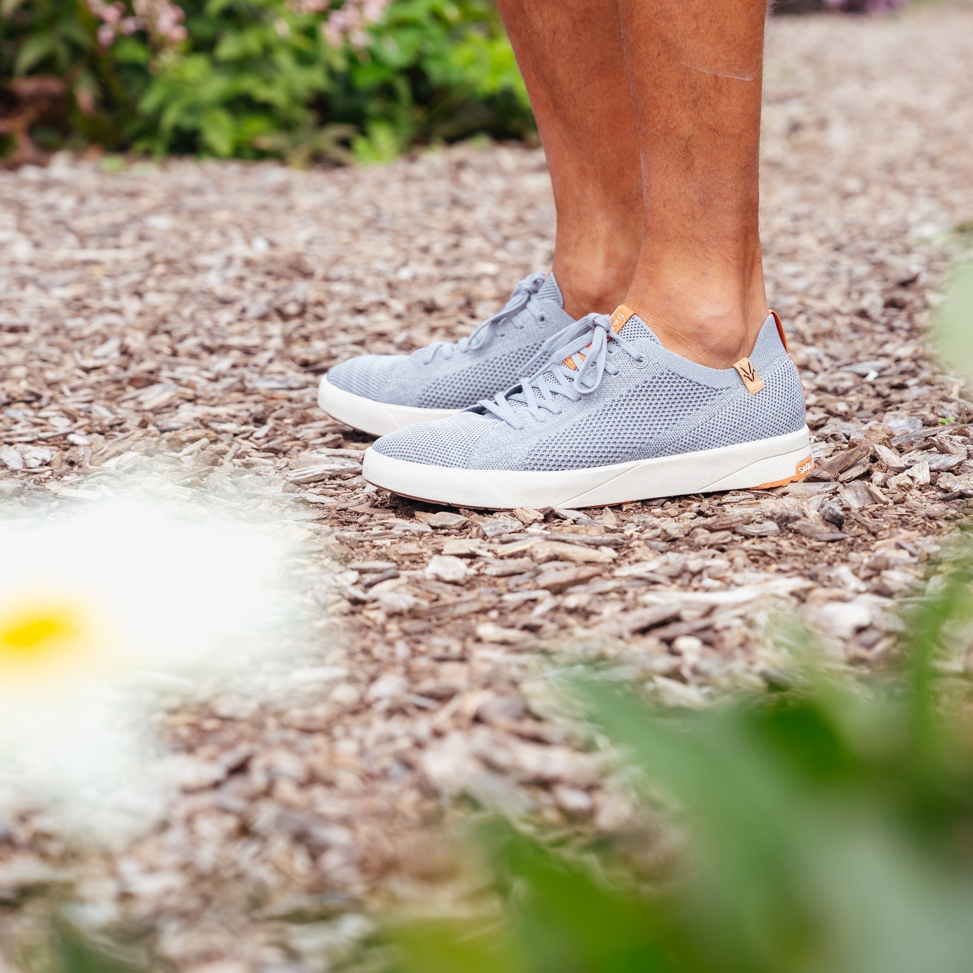 A person wearing light gray knit sneakers with white soles, standing on a gravel path outdoors with blurred greenery in the foreground and background.