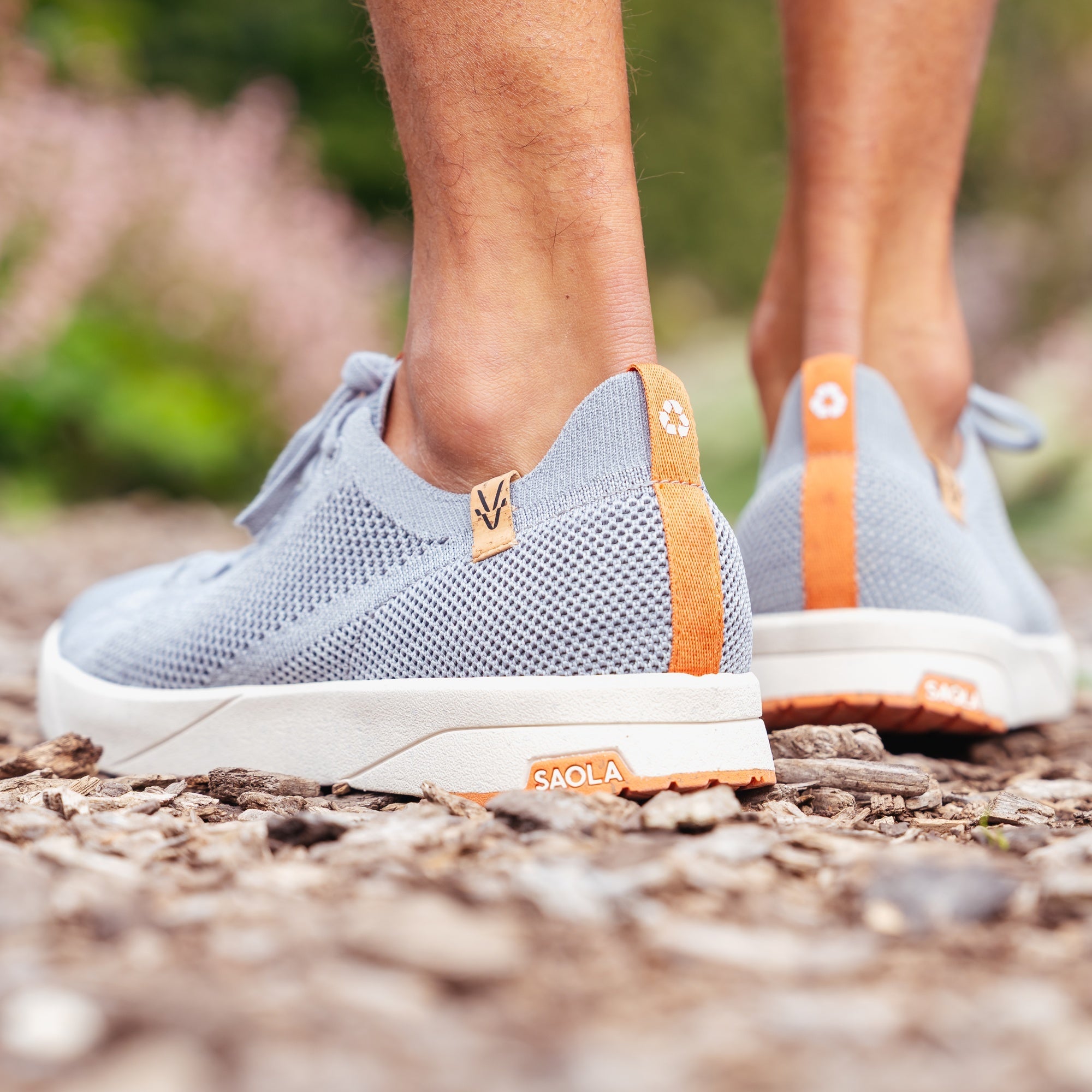 A close-up of a person's feet wearing light gray knit slip-on sneakers with orange heel tabs and white soles, standing on a gravel path outdoors. Visible branding includes 'SAOLA' on the shoe and a monogram logo on the heel tab.