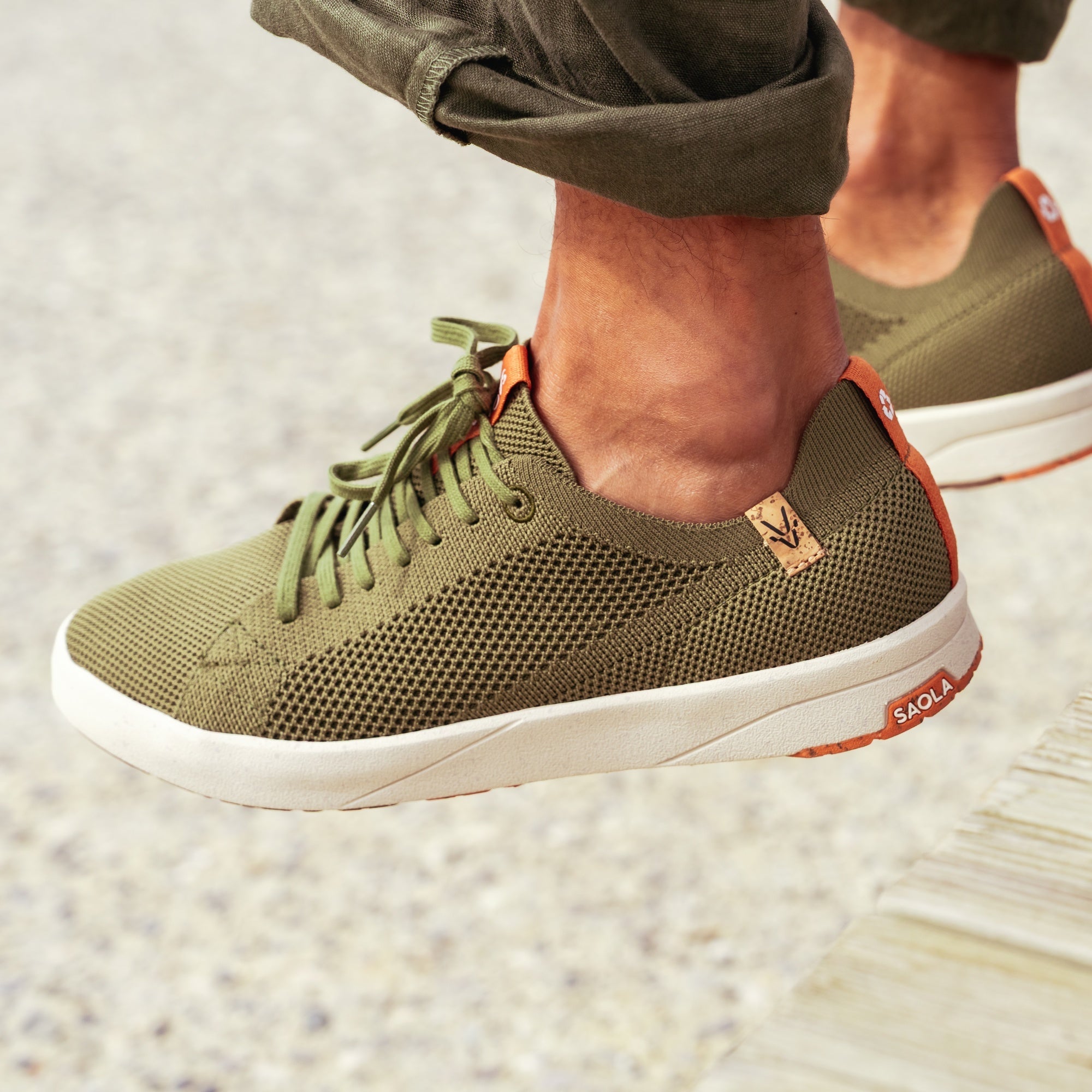 A close-up of a person wearing olive green mesh sneakers with white soles and orange accents, standing on a light concrete surface.