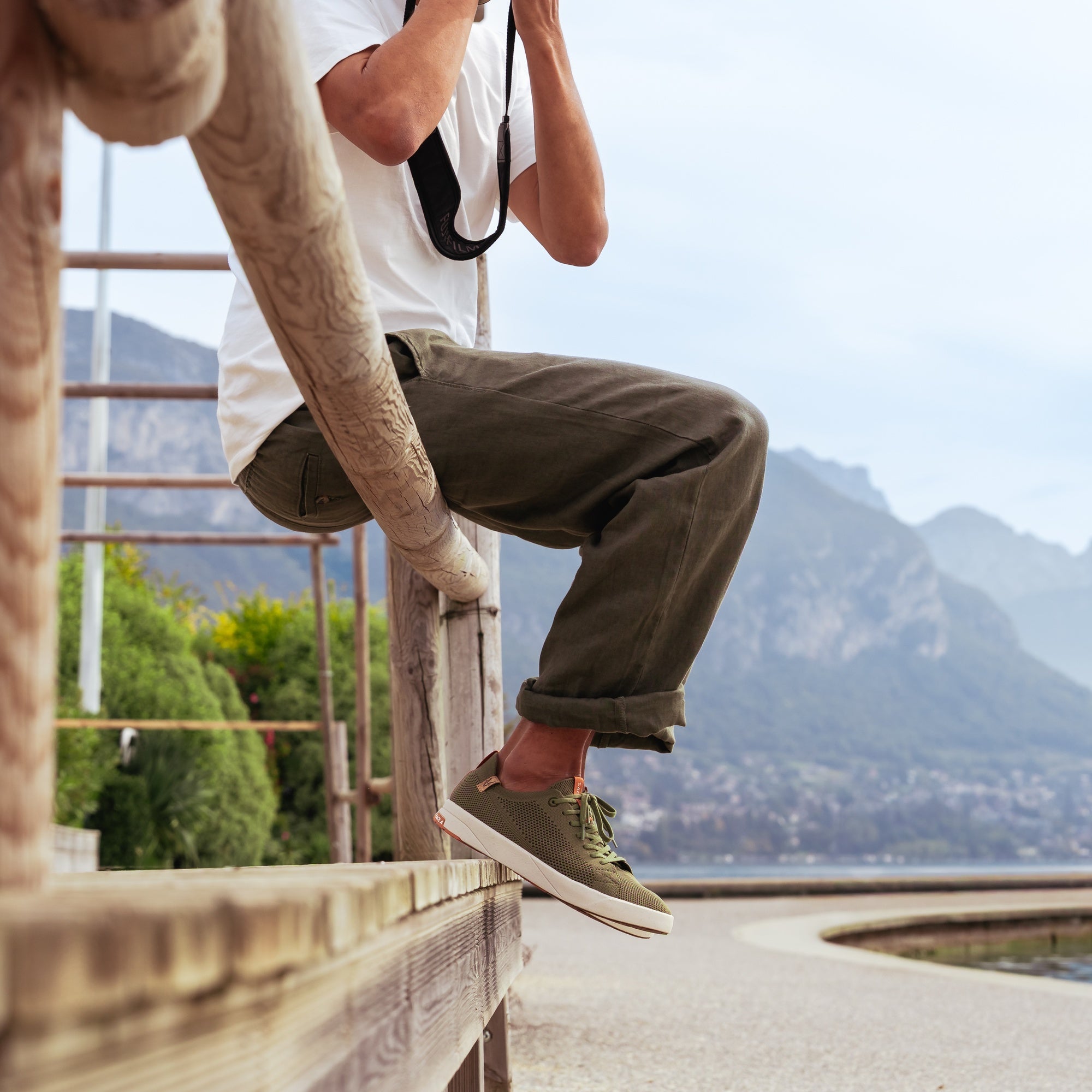 A person sitting on a wooden railing outdoors, wearing olive green trousers and matching low-top sneakers with a white t-shirt. The background shows a scenic mountain and lake view.