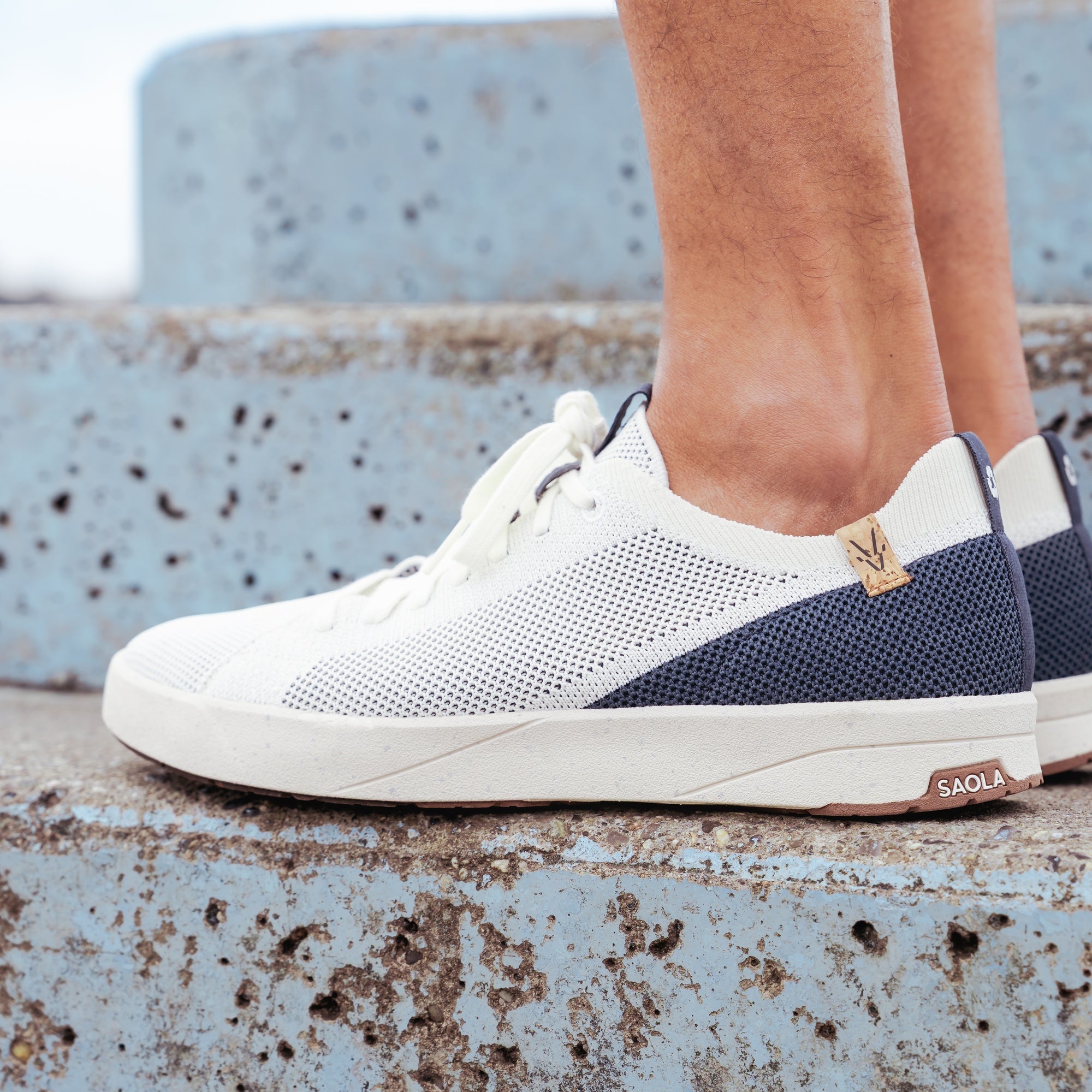 White and navy blue knit sneakers worn on a person's feet, standing on a weathered blue concrete ledge. The shoe features a white knit upper, navy blue mesh side panels, white laces, and a small gold logo on the heel.