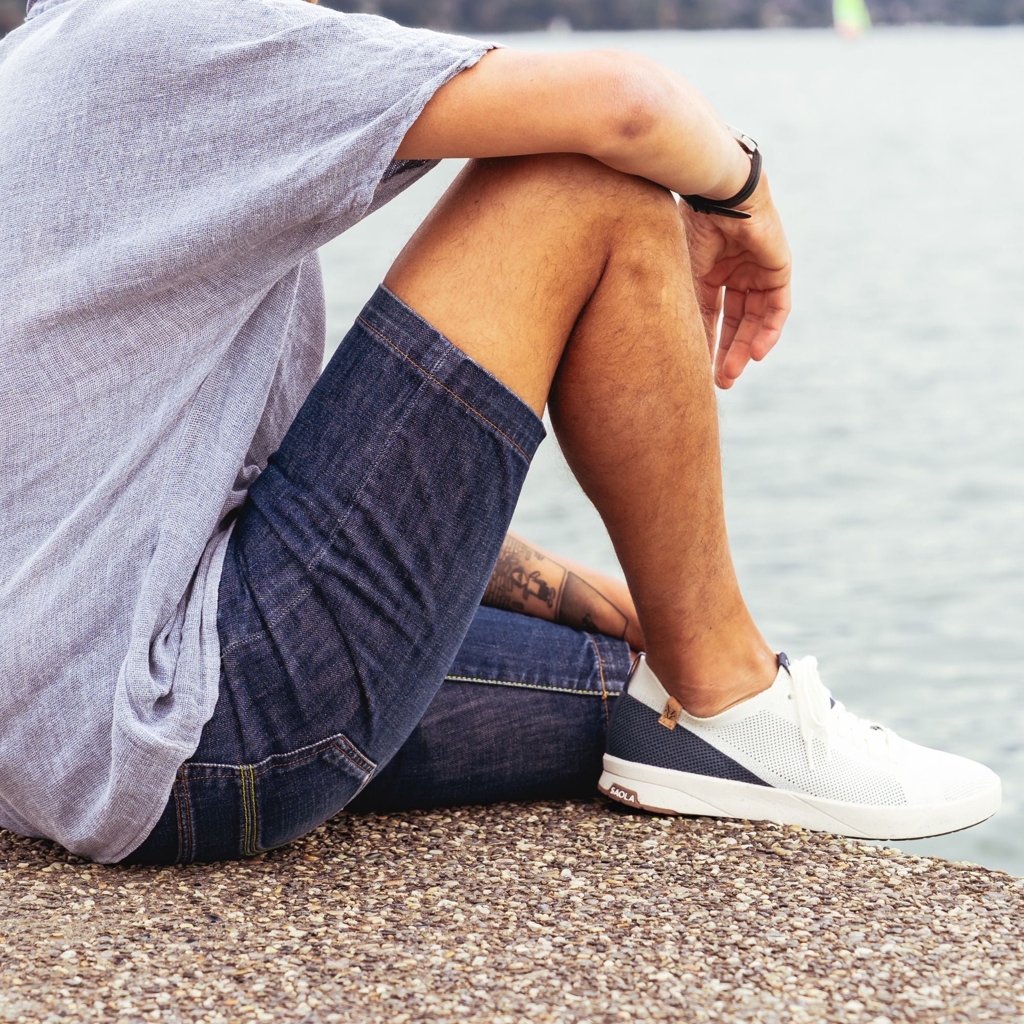 A person sitting on a pebble shore by the water, wearing a light gray oversized T-shirt, dark denim shorts, and white slip-on shoes with navy accents.