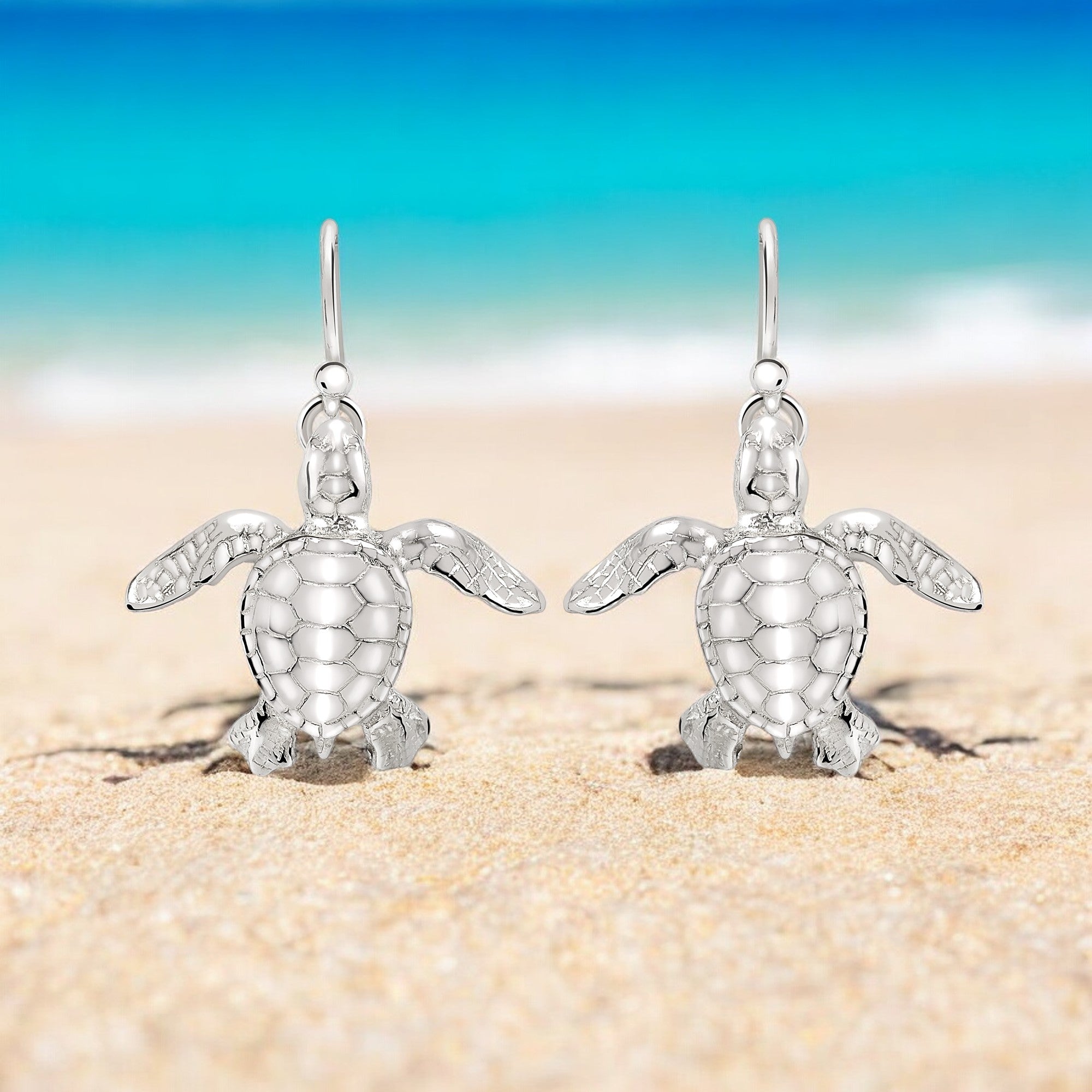 Silver turtle earrings on a sandy beach with a blue sky background
